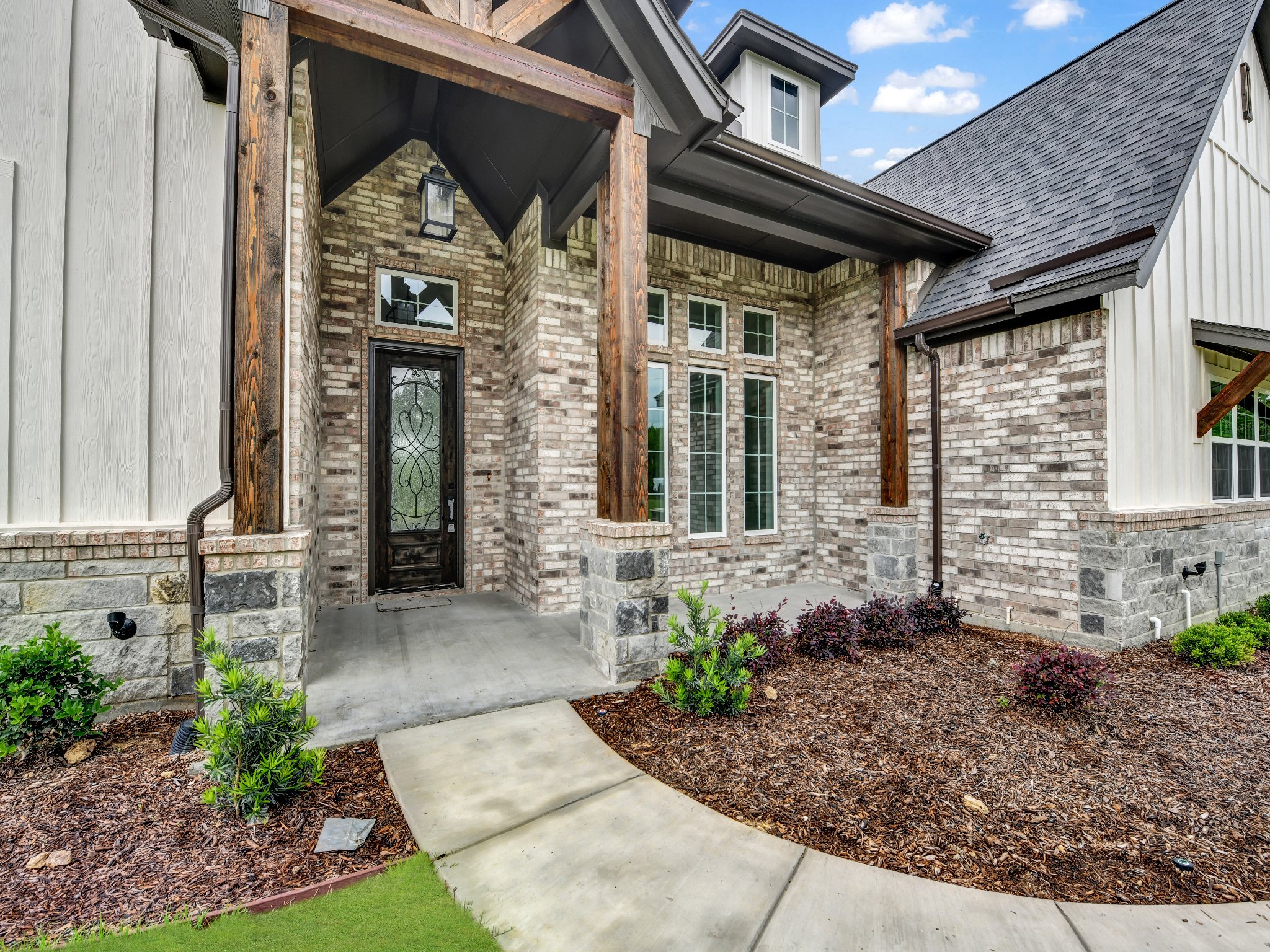 Entrance of a house with brick and siding exterior, stone foundation, black front door with decorative glass, and a concrete pathway leading to the door. There are plants and mulch in the landscaping.