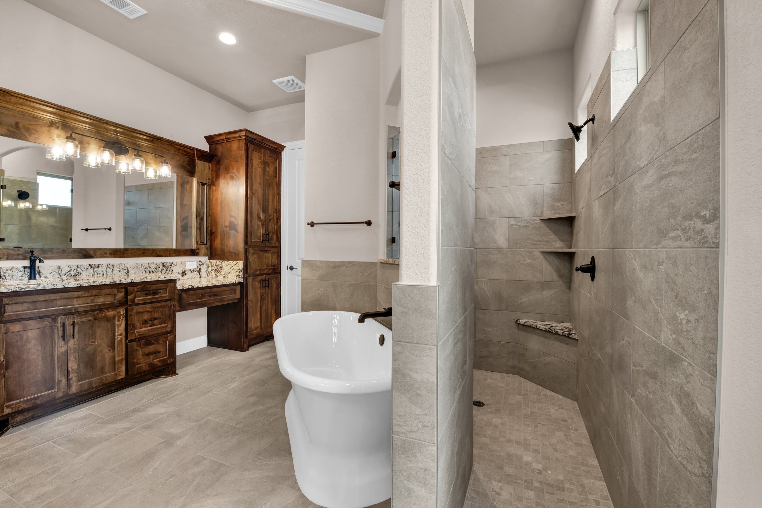 Bathroom with a large mirror, wooden cabinets, granite countertop, white bathtub, and a walk-in shower with beige tiles and built-in shelves.