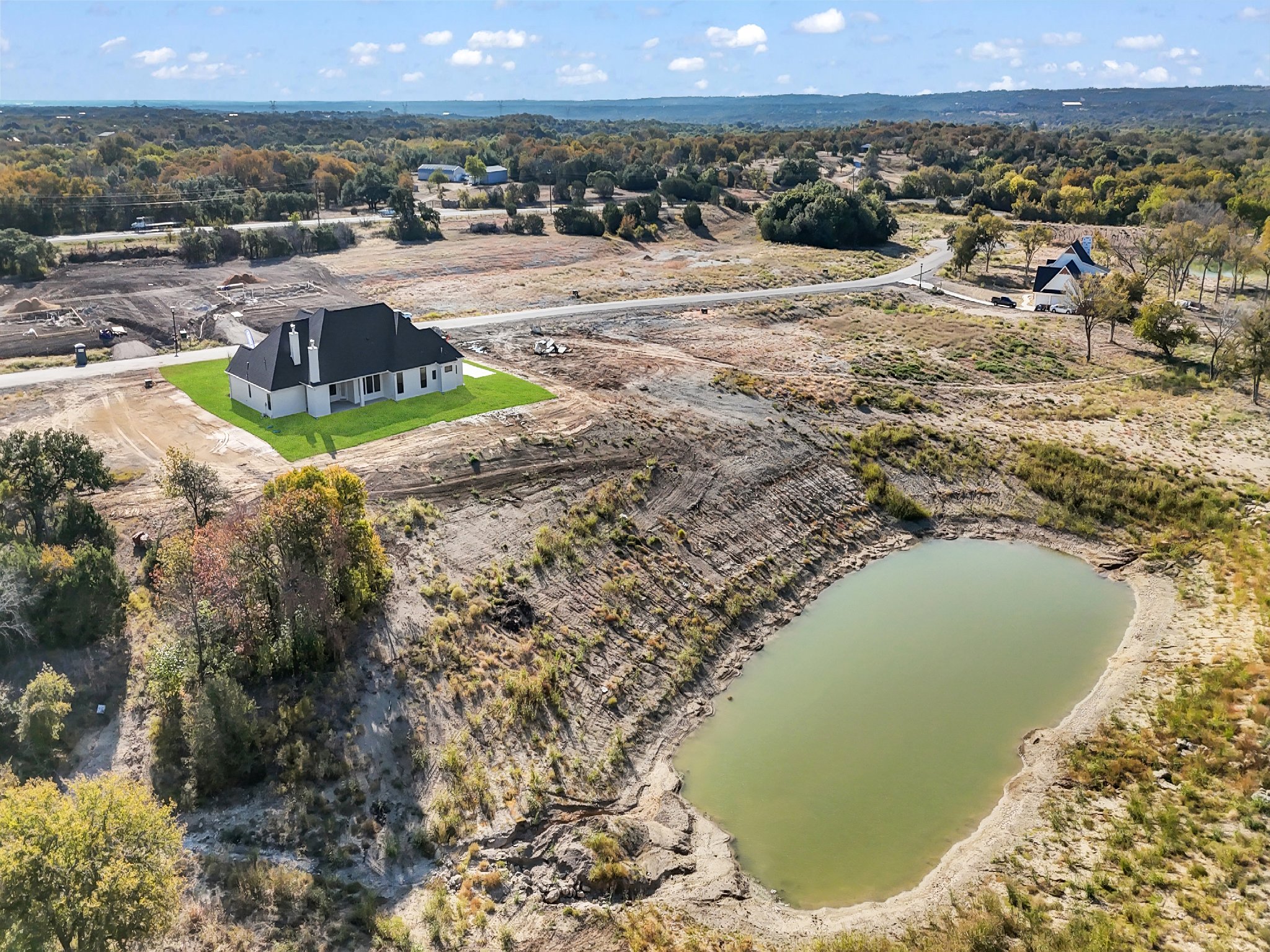 A house under construction on a grassy lot next to a small pond, in a semi-rural area with trees, a road, and other homes nearby, under a partly cloudy sky.