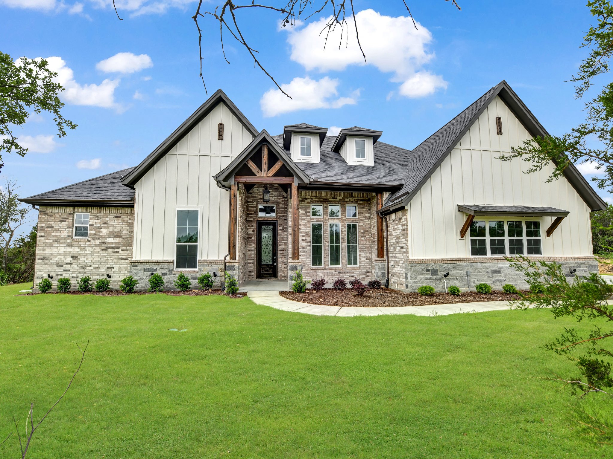 A modern house with a mix of brick, stone, and vertical siding, featuring multiple pitched roofs, windows, and a front door with a decorative glass panel, surrounded by a well-maintained lawn and landscaped garden.