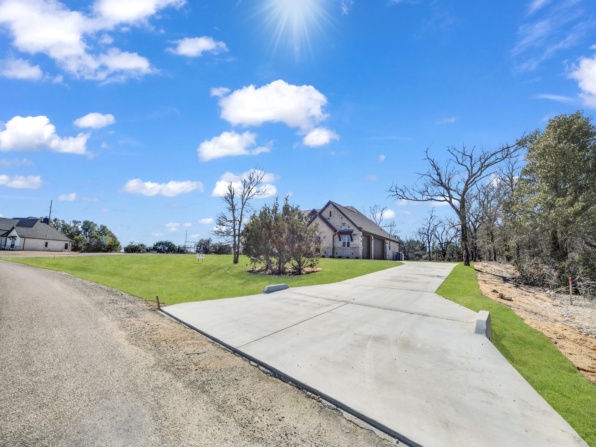 Newly paved driveway leading to a house with a gray roof, surrounded by grass, leafless trees, and a clear blue sky with scattered clouds and bright sunlight.