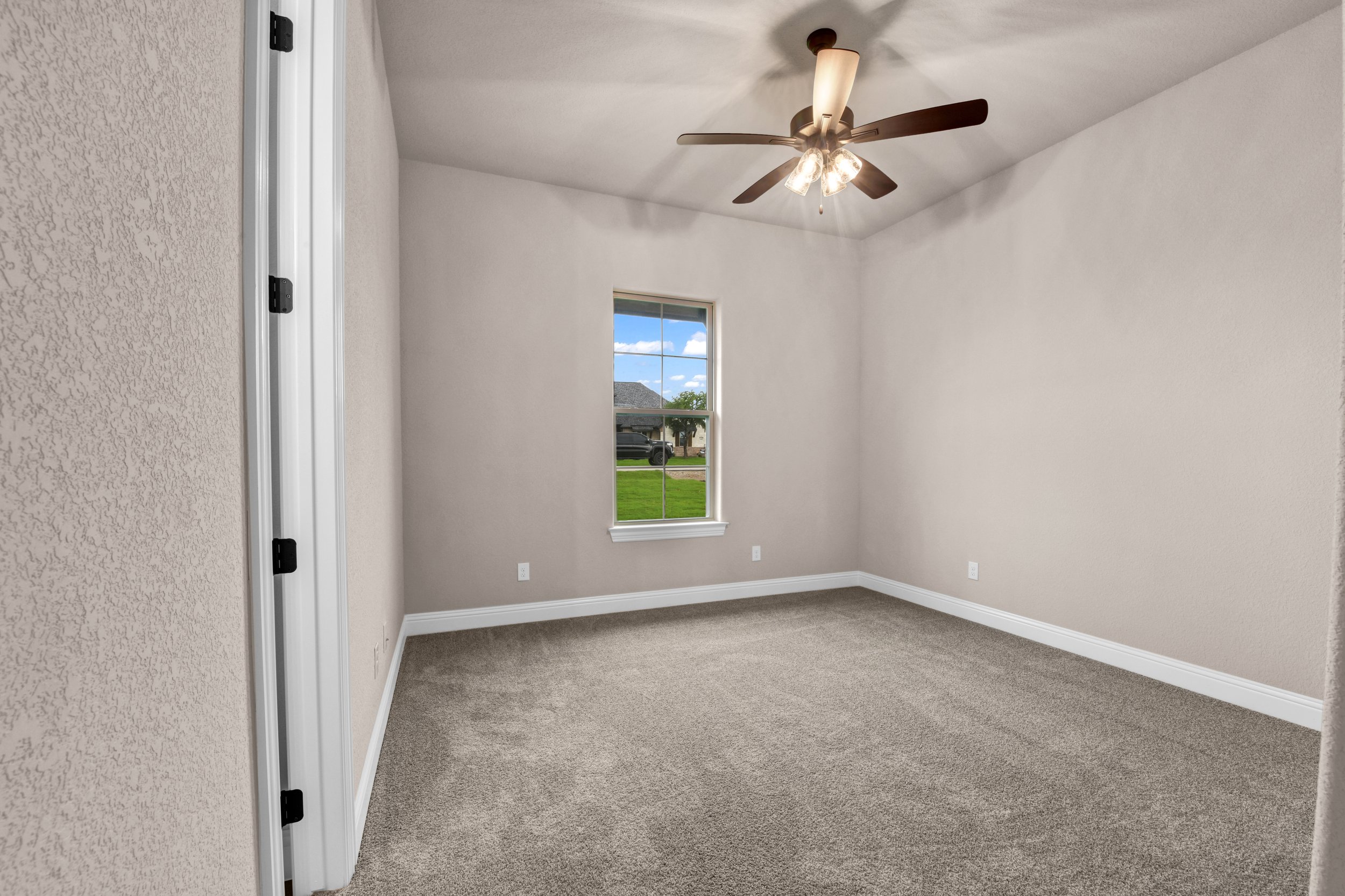 Empty room with beige walls, a white window showing a yard outside, ceiling fan with lights, and carpeted flooring.