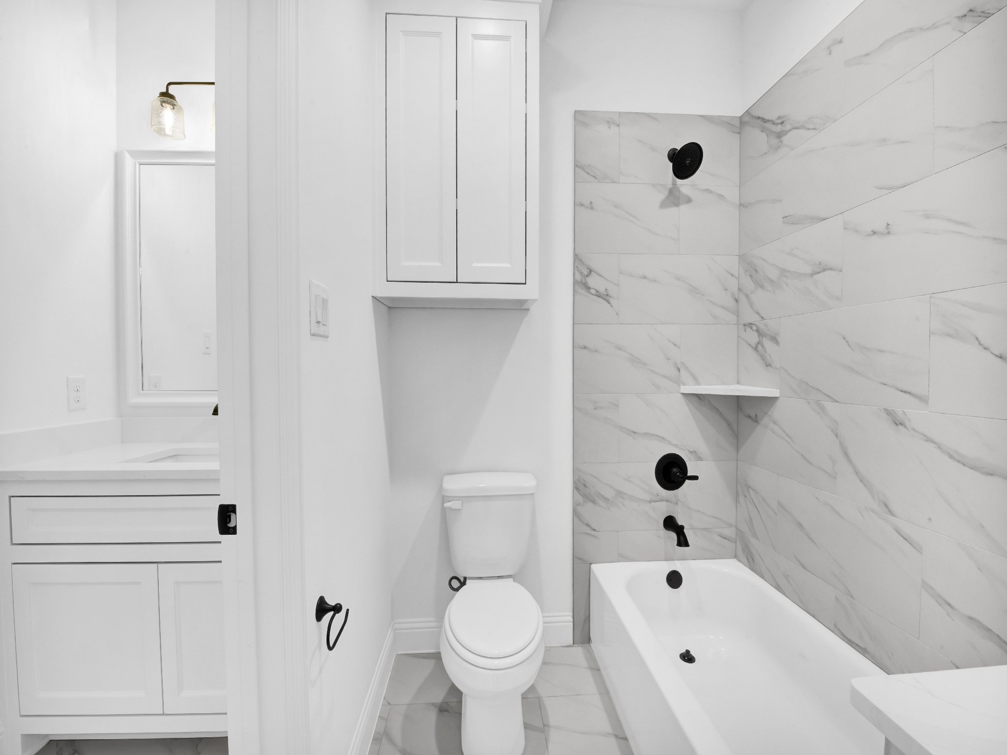 A modern white bathroom with a toilet, a marble-tiled bathtub with black fixtures, and a white cabinet. There is a wall-mounted light fixture with a glass shade and a mirror with a frame in the background.