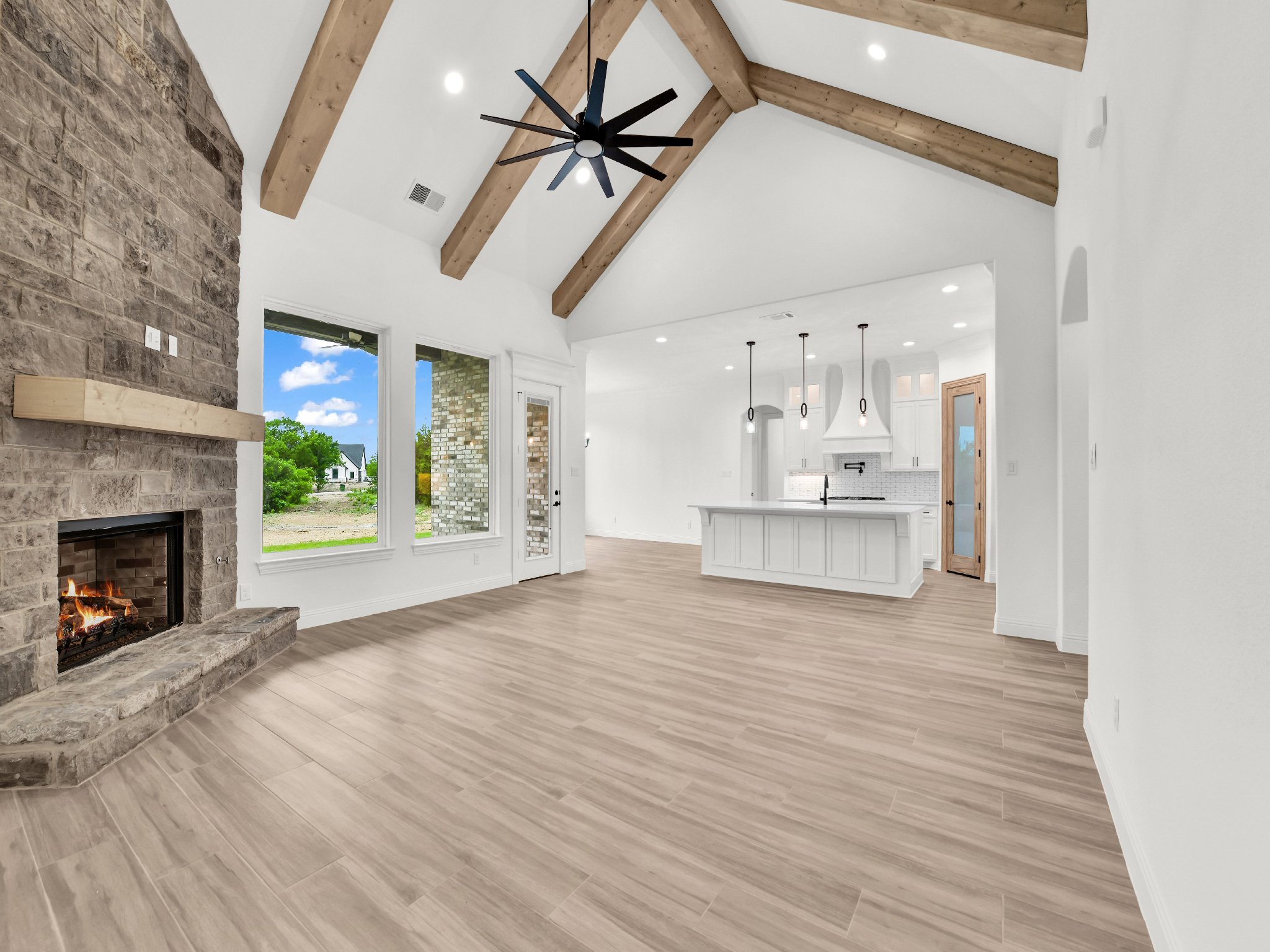 Open concept living room and kitchen with vaulted ceilings, exposed wooden beams, a black ceiling fan, a stone fireplace, large windows, and a white kitchen island.