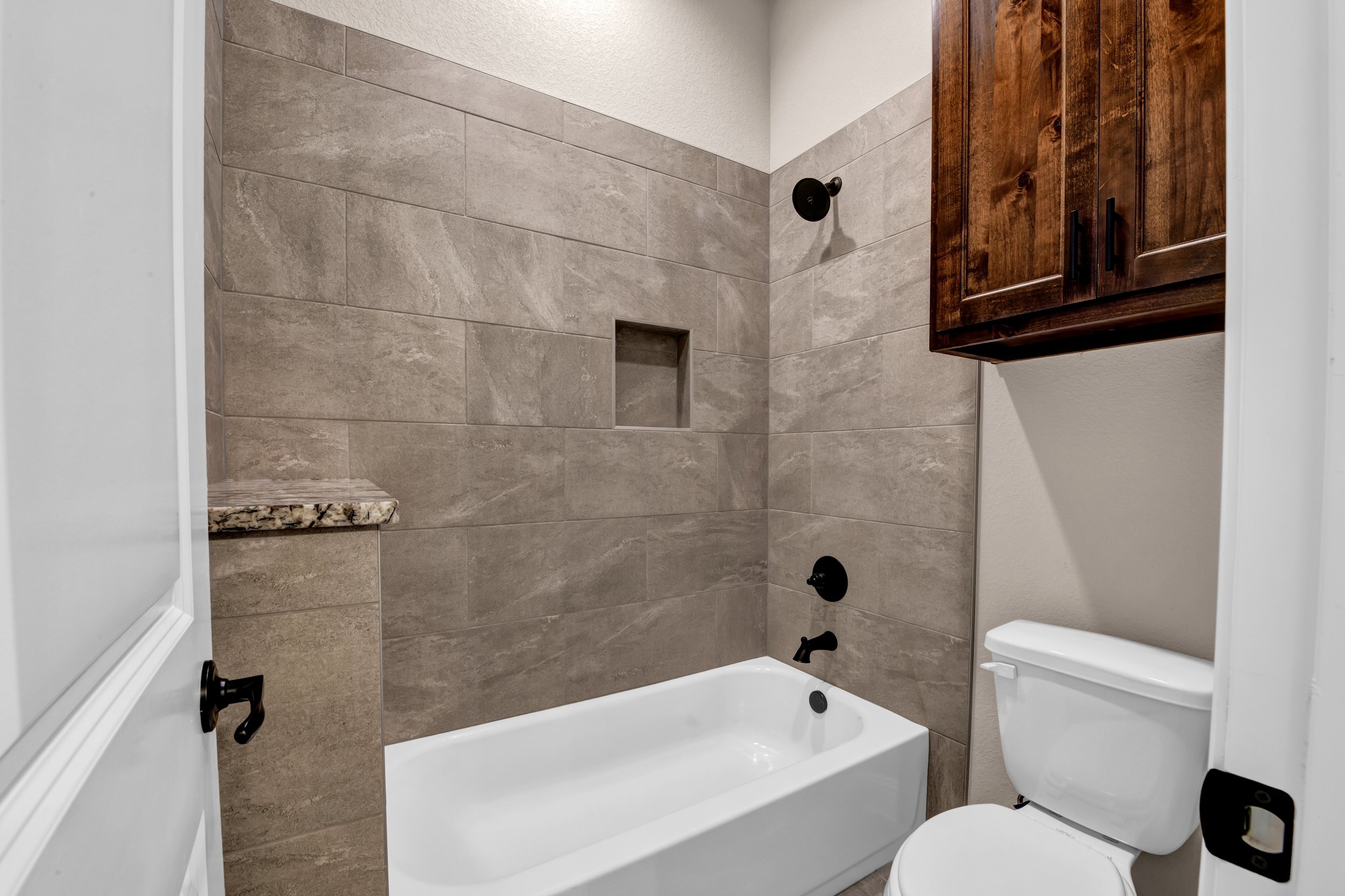 Bathroom with a white bathtub, beige tiled walls, a wooden cabinet above the toilet, and black fixtures including a showerhead, control knob, and faucet.