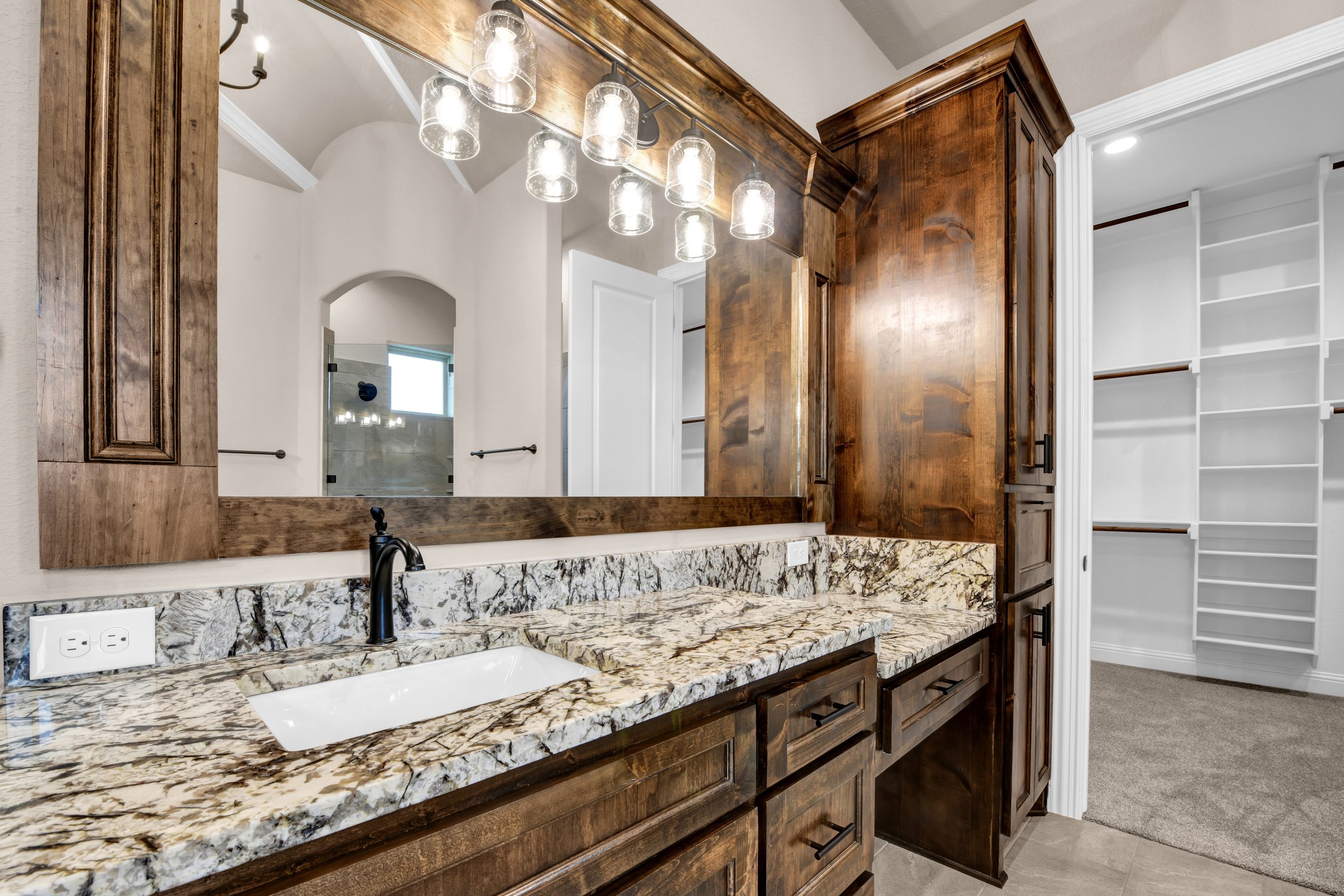Bathroom vanity with a granite countertop, wooden cabinets, a large mirror, and a black faucet. There are light fixtures with glass shades above the mirror, and a walk-in closet with white shelving in the background.