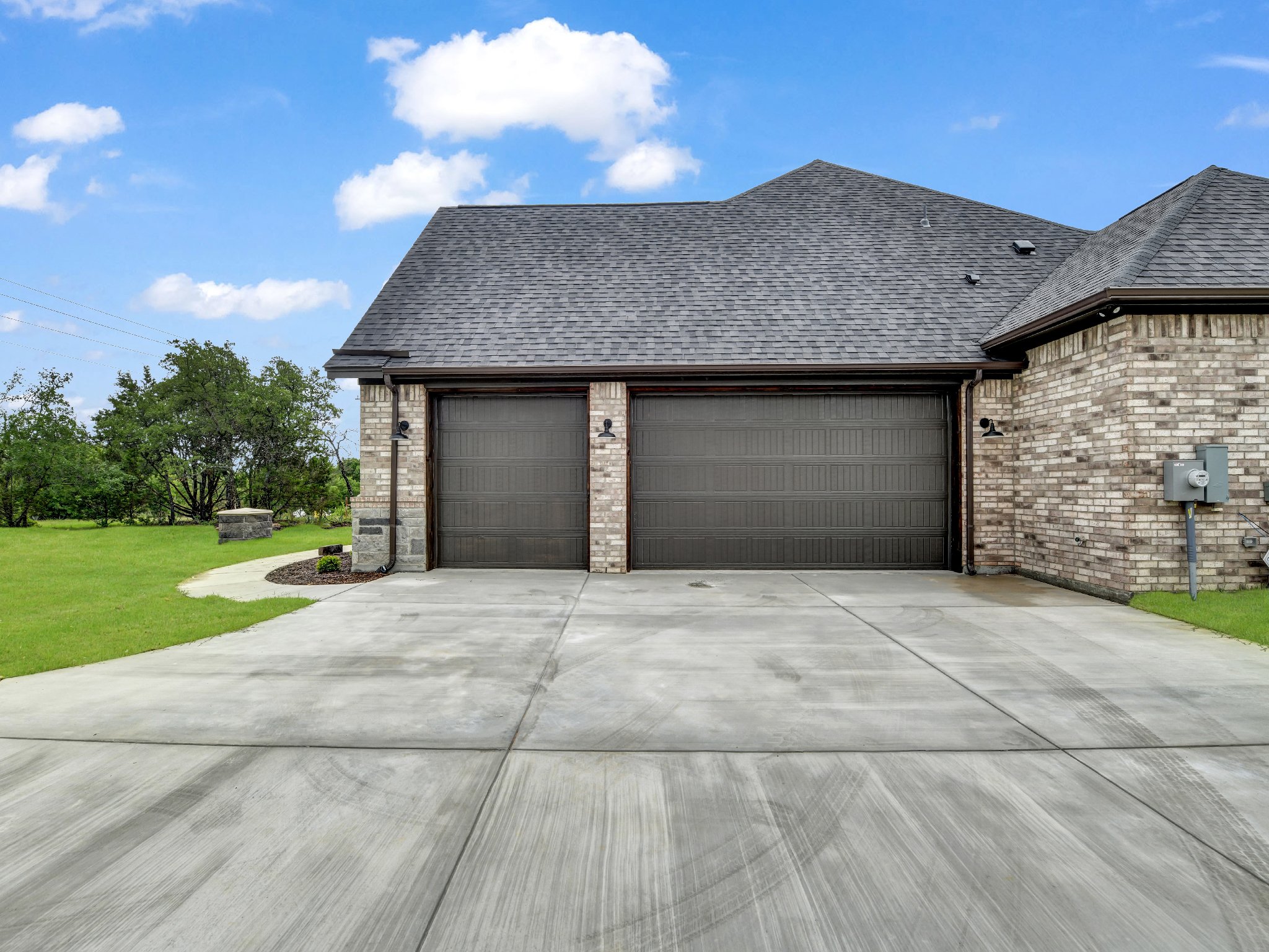 Front view of a house with a brick exterior, gray garage doors, a concrete driveway, and a grassy yard with trees in the background under a blue sky with clouds.