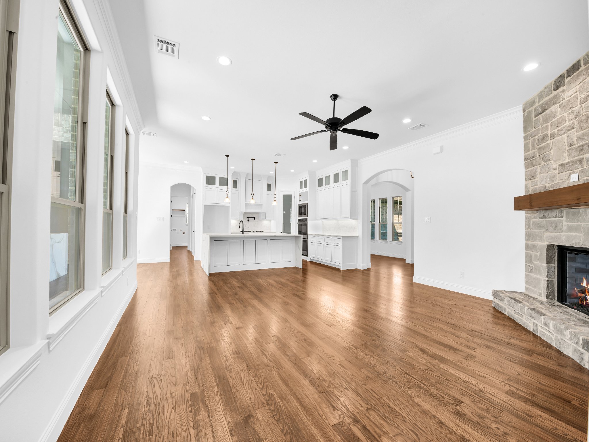 Empty living room with large windows, hardwood floors, white walls, a black ceiling fan, and a stone fireplace.
