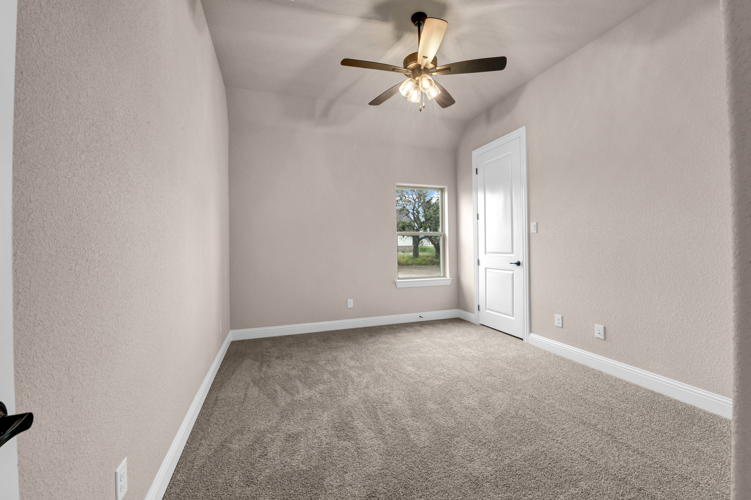 Empty off-white room with beige carpet, a window showing trees outside, closed white door, ceiling fan with light, electrical outlets, and baseboards.
