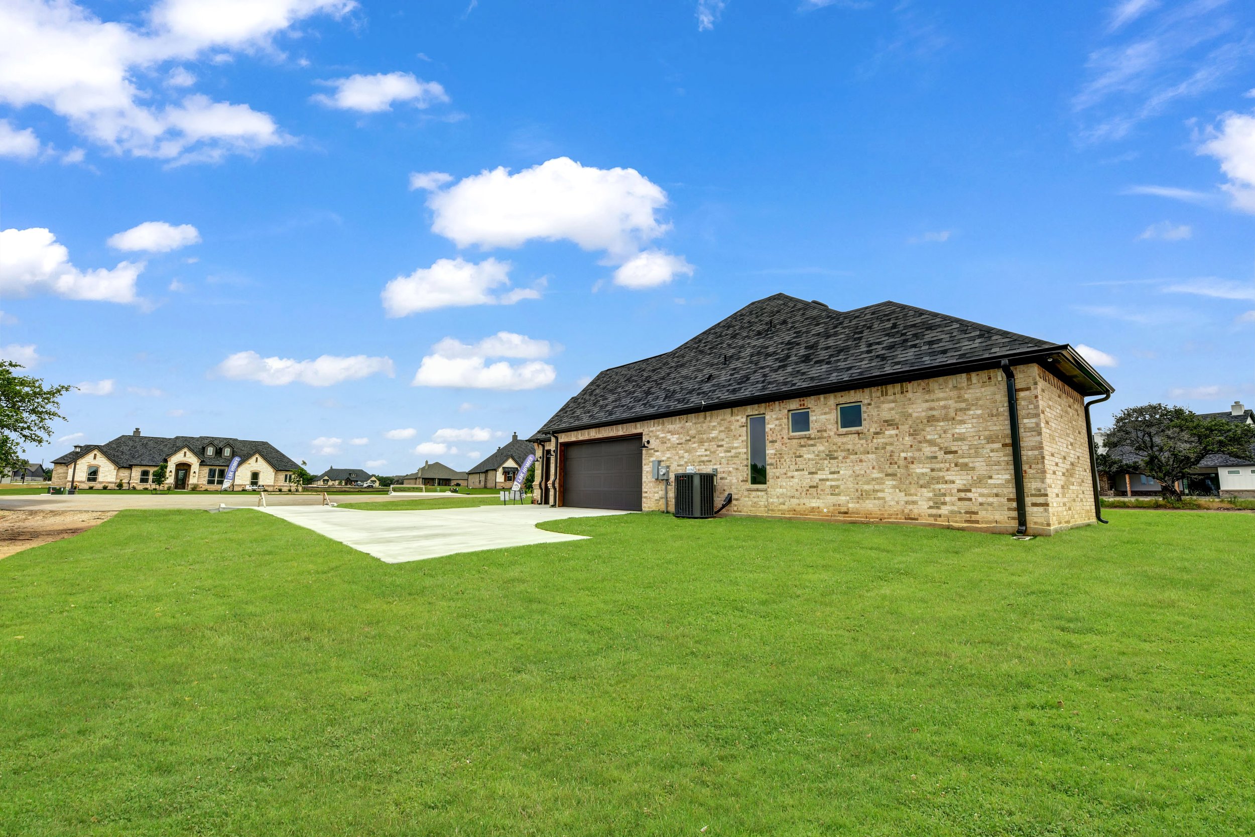 A brick house with a dark shingled roof, attached garage, and an air conditioning unit on a green lawn under a blue sky with scattered clouds.