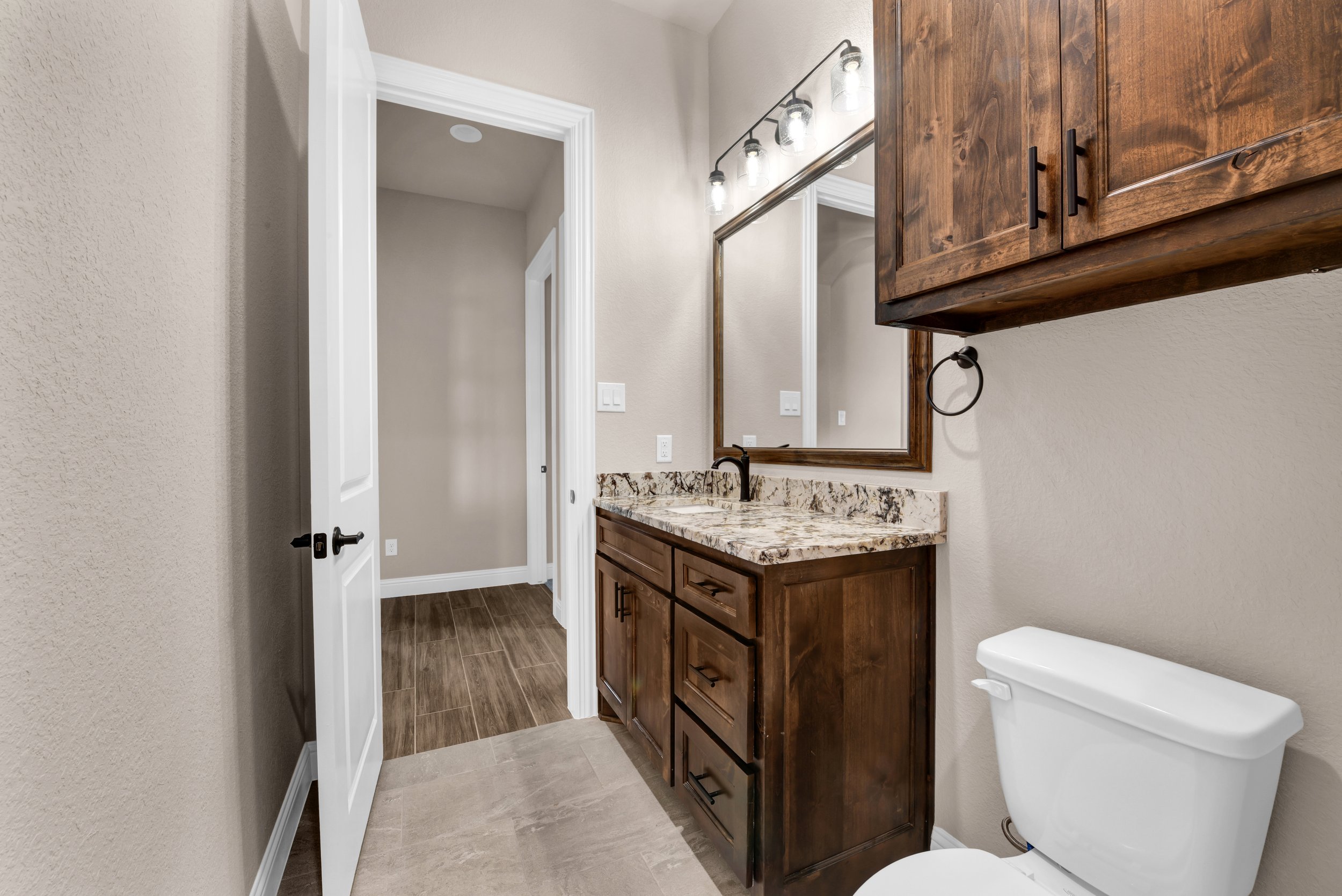 Bathroom with beige walls, wooden vanity with granite countertop, large mirror, wall-mounted light fixture, white toilet, and wooden cabinets above the vanity.