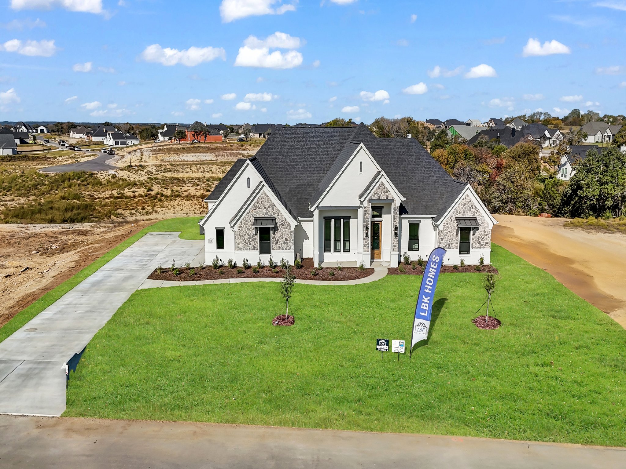Newly constructed modern house with white exterior, stone accents, black roof, and a well-maintained green lawn. A blue LBK Homes flag is in the front yard, with a concrete driveway on the left side leading to the house. Surrounding area shows partly