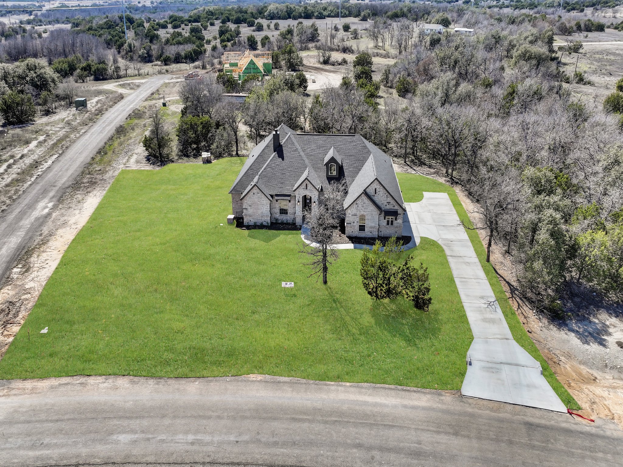 An aerial view of a newly constructed house with a gray roof, situated on a large grassy lot with a concrete driveway, surrounded by a mix of bare and green trees, with other development in progress in the background.