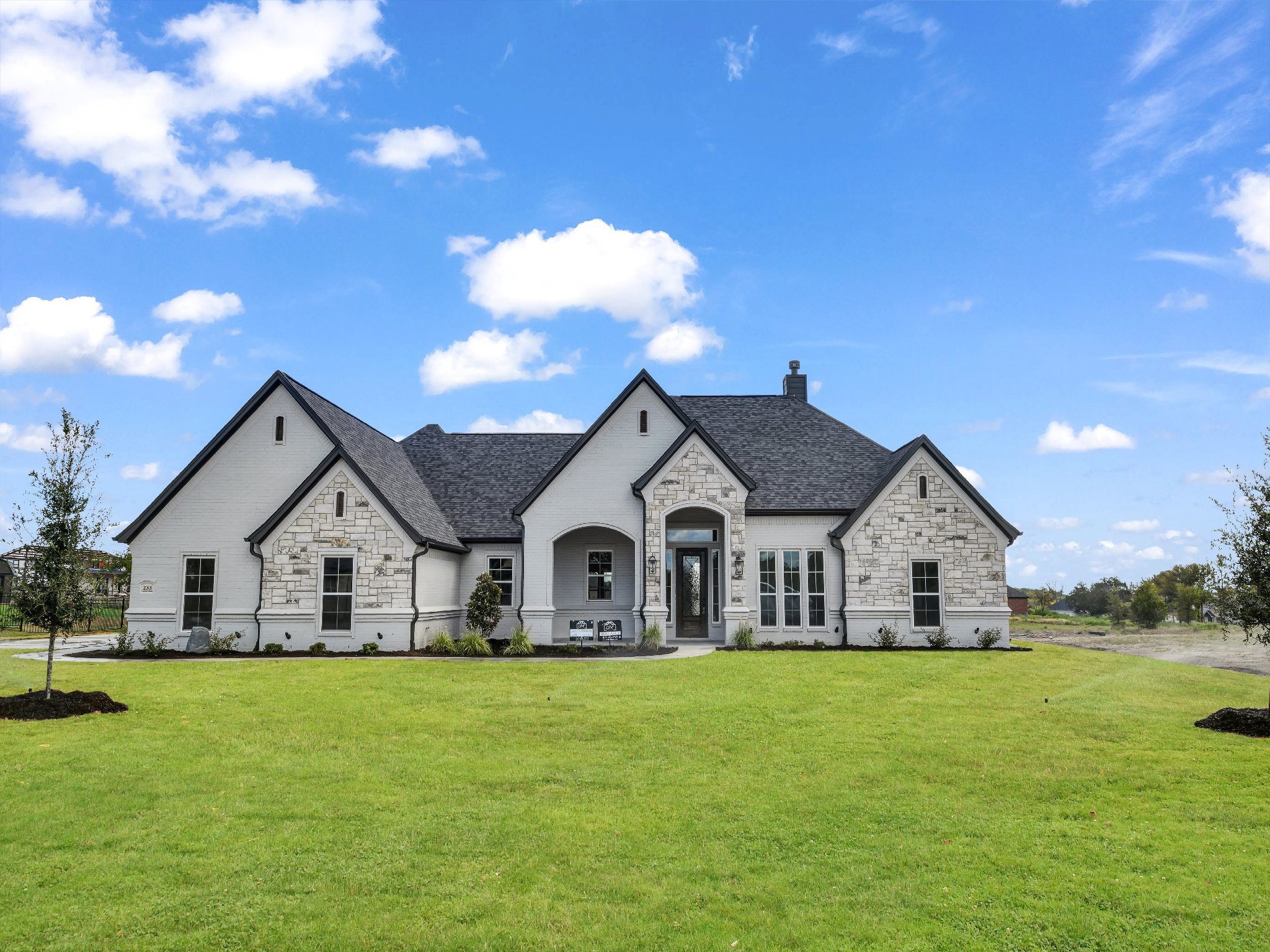 Large modern house with white brick and stone exterior, black roof, front yard with green grass, small trees, and blue sky with clouds.