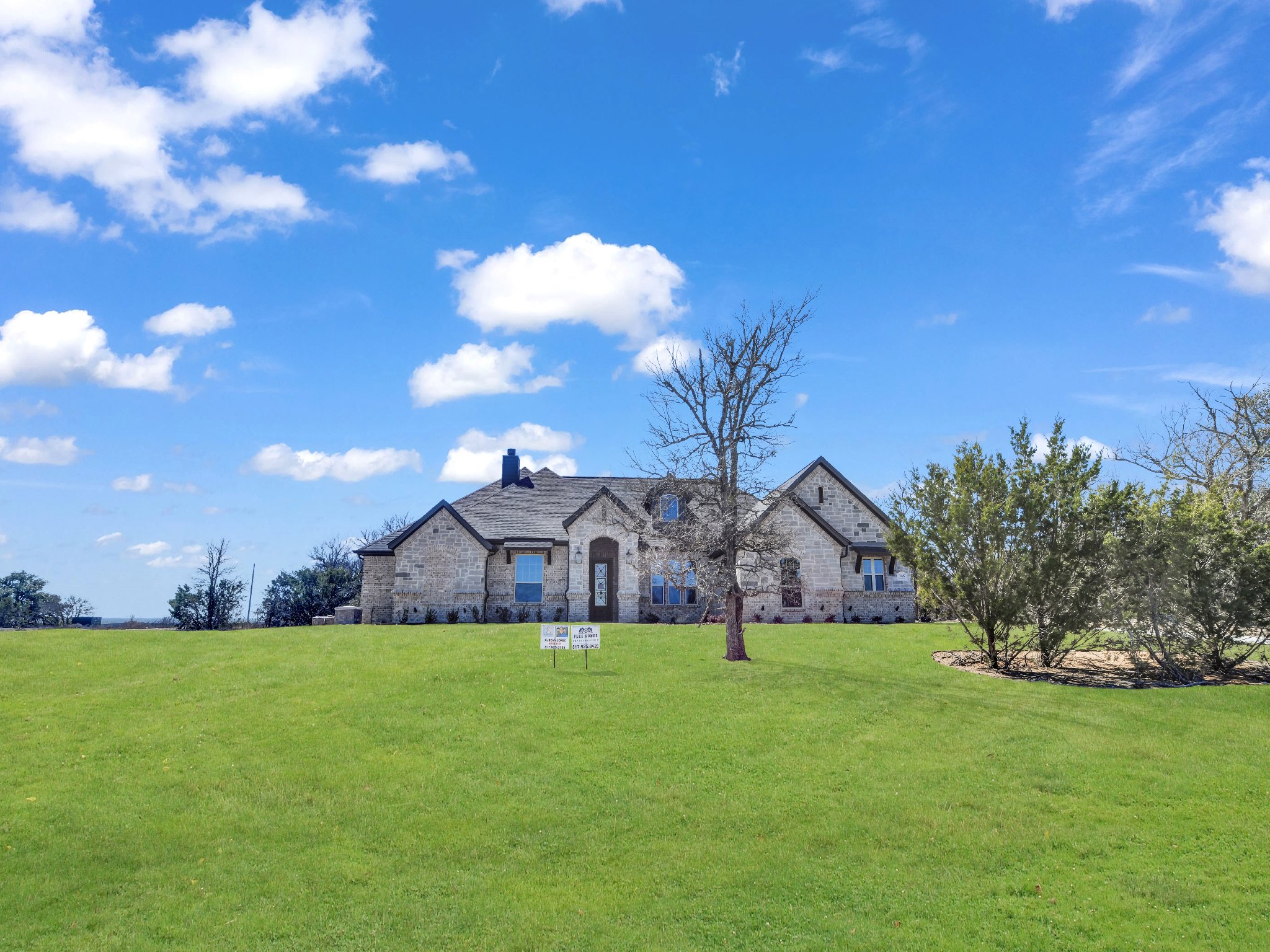 A large brick house with multiple windows and a chimney, situated on a lush green lawn with some trees and shrubs, under a bright blue sky with scattered clouds.