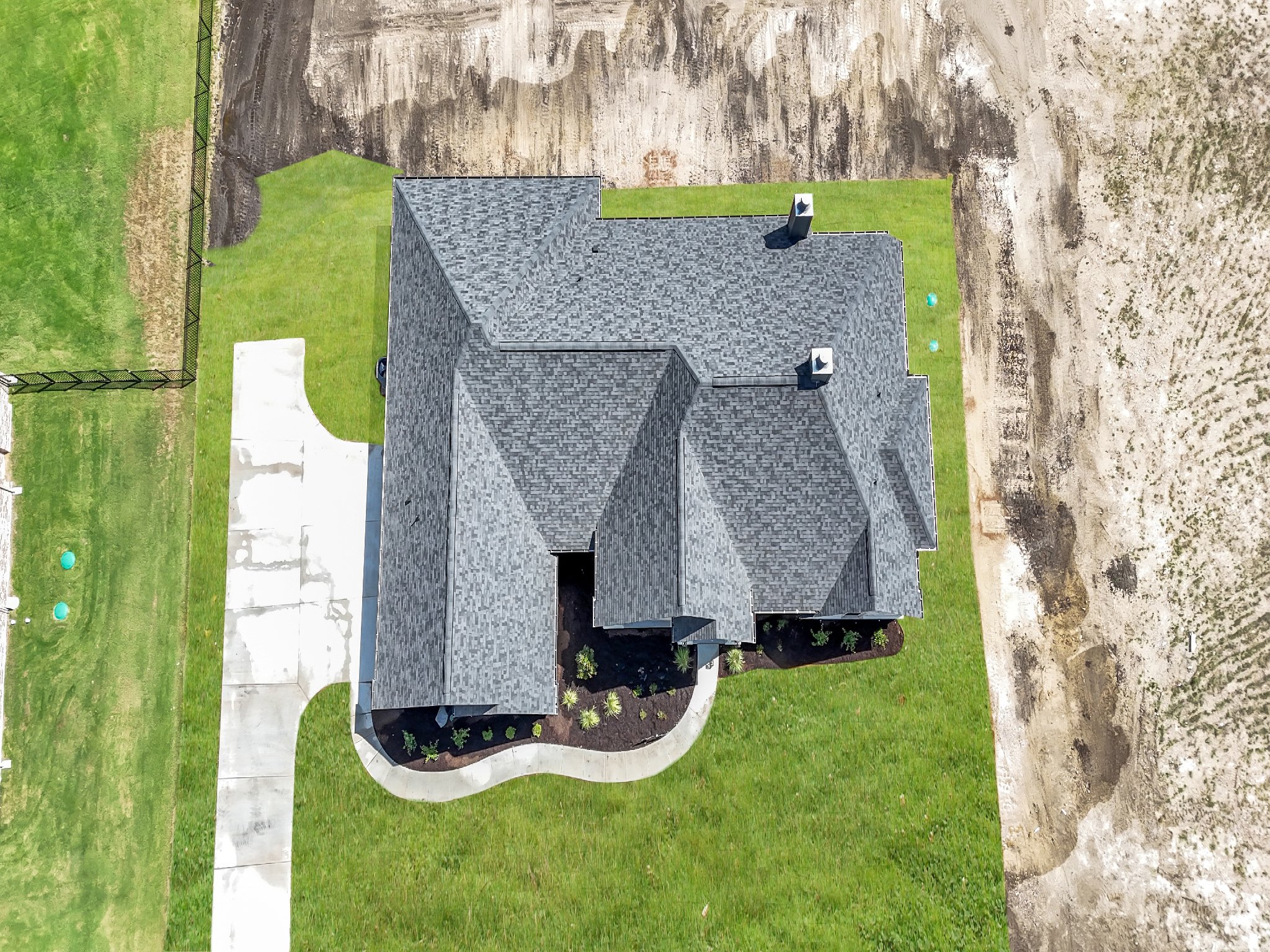 Aerial view of a house with a gray shingle roof, surrounded by green grass and a concrete driveway, with a fenced yard and adjacent dirt area.