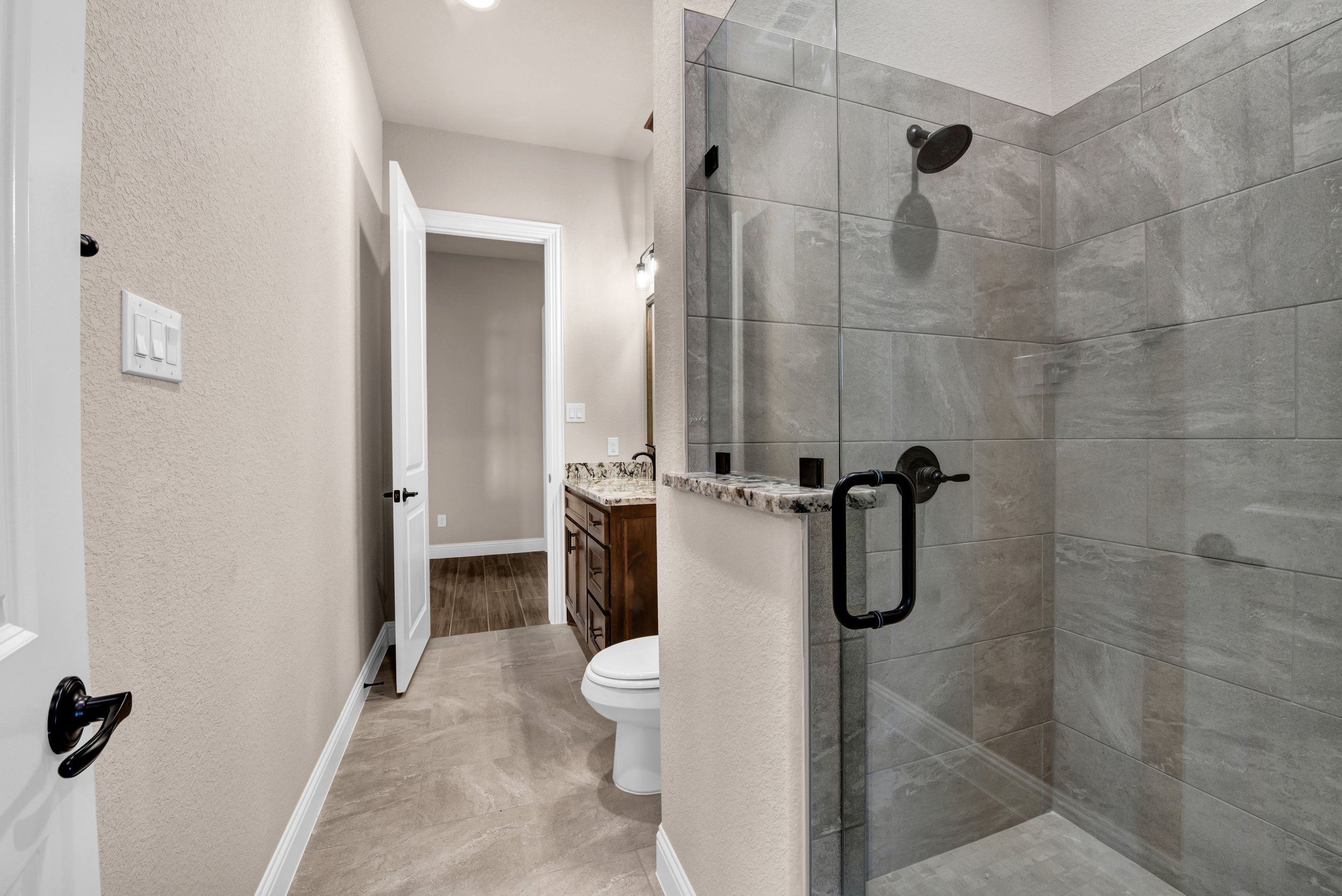 Bathroom with a walk-in shower with gray tile and a glass door, a toilet, a vanity with a granite countertop, and wood flooring.