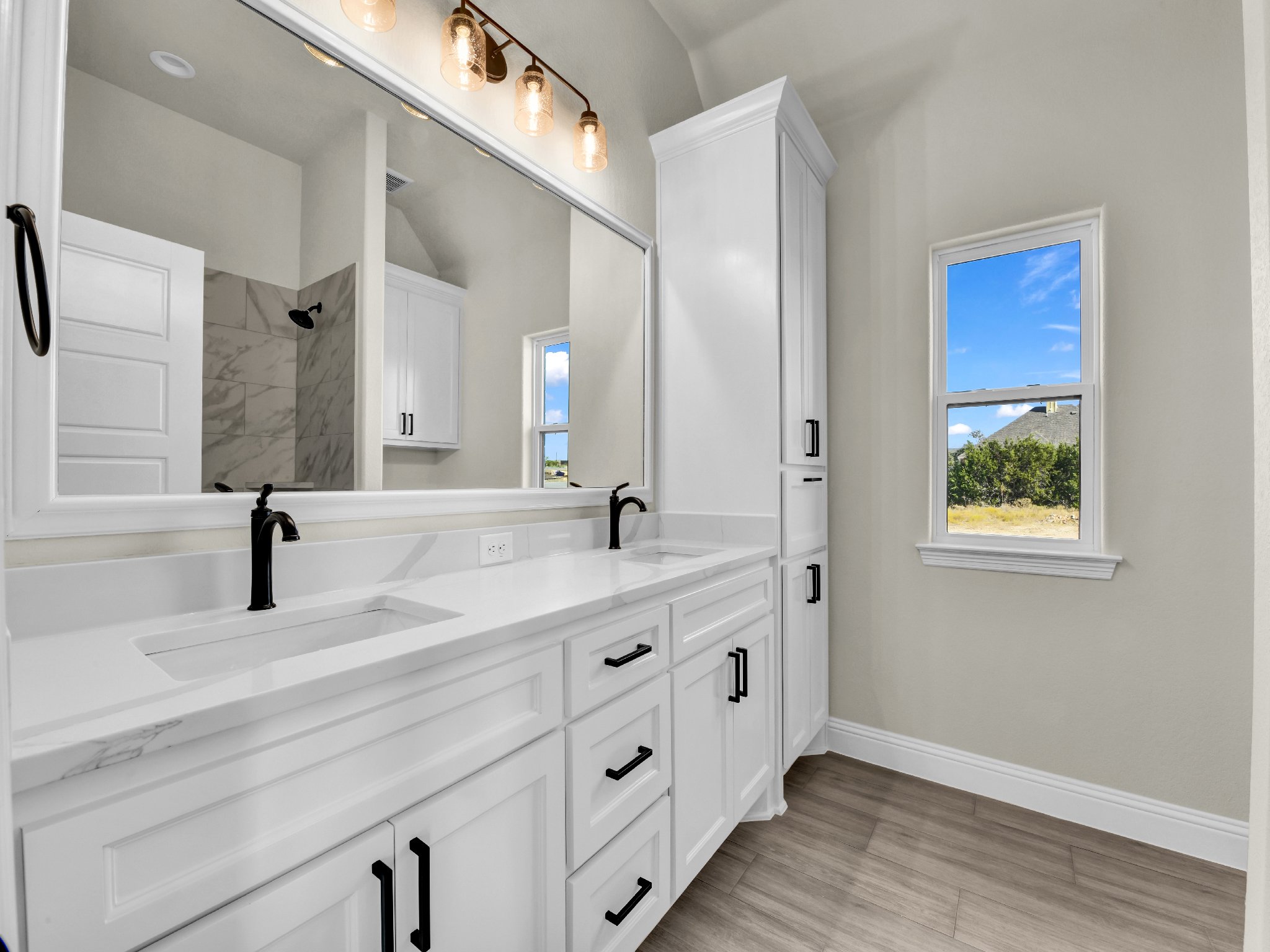 Clean modern white bathroom vanity with dual sinks, black faucets, large mirror, and two windows showing outdoor scenery.