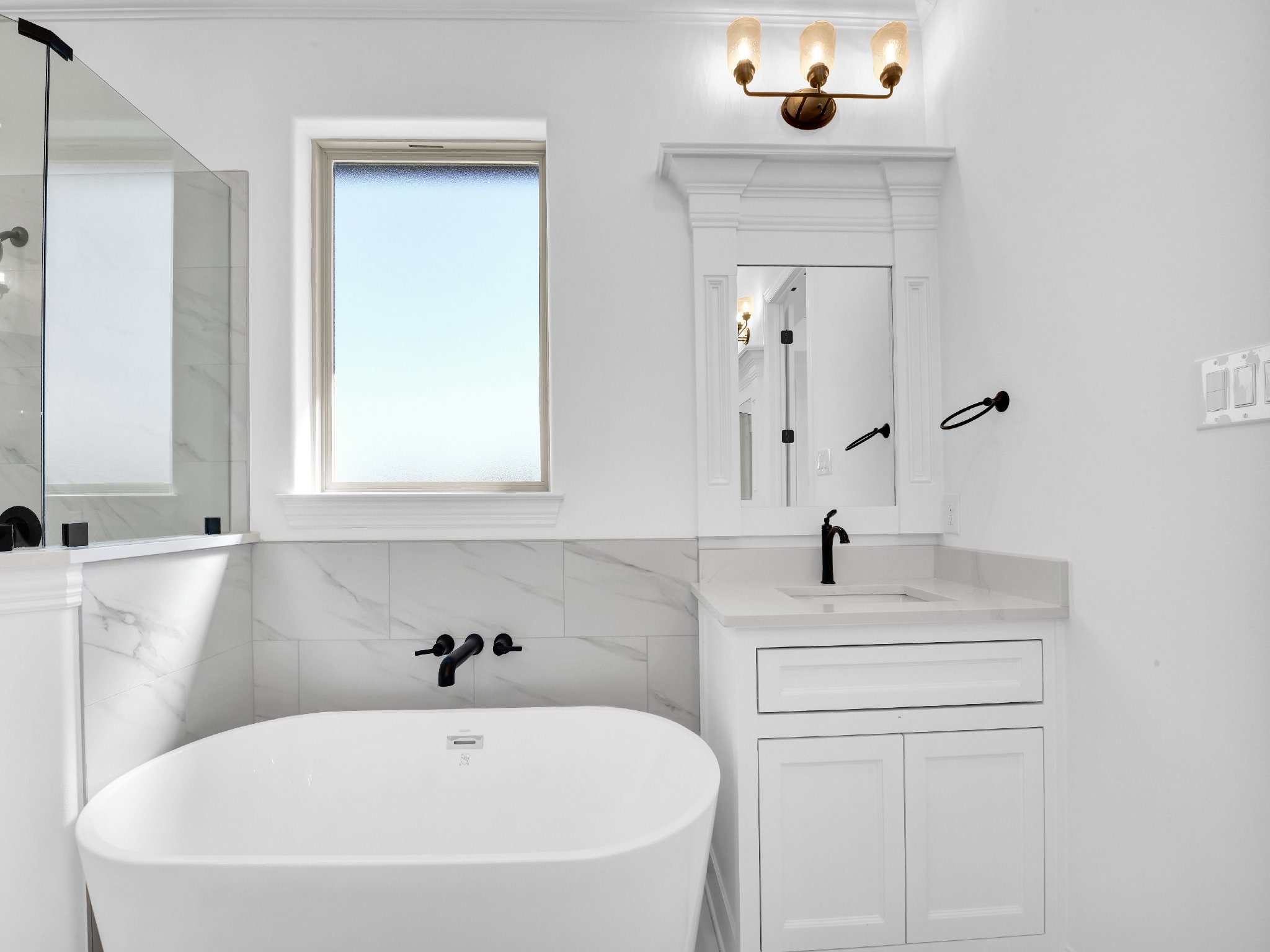 Modern white bathroom with a bathtub, window, mirror, black fixtures, and a light fixture with three bulbs.