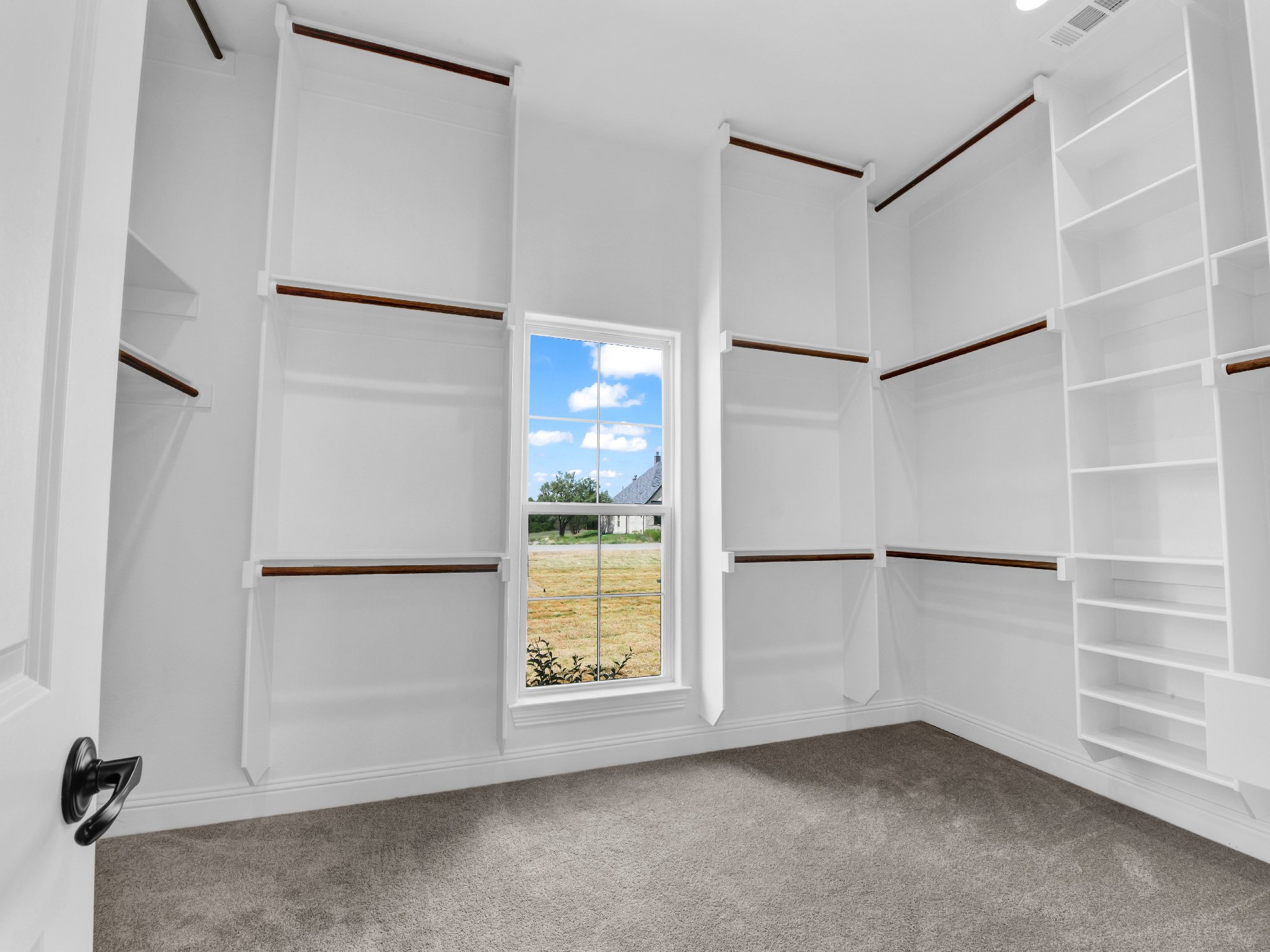 Empty walk-in closet with white walls, built-in shelves and hanging rods, and a window showing a grassy yard with a house in the distance.