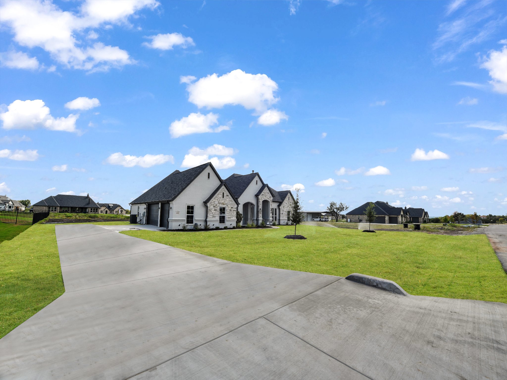 A modern house with white exterior walls and black roofing, situated on a green lawn with a curved concrete driveway under a blue sky with scattered clouds.