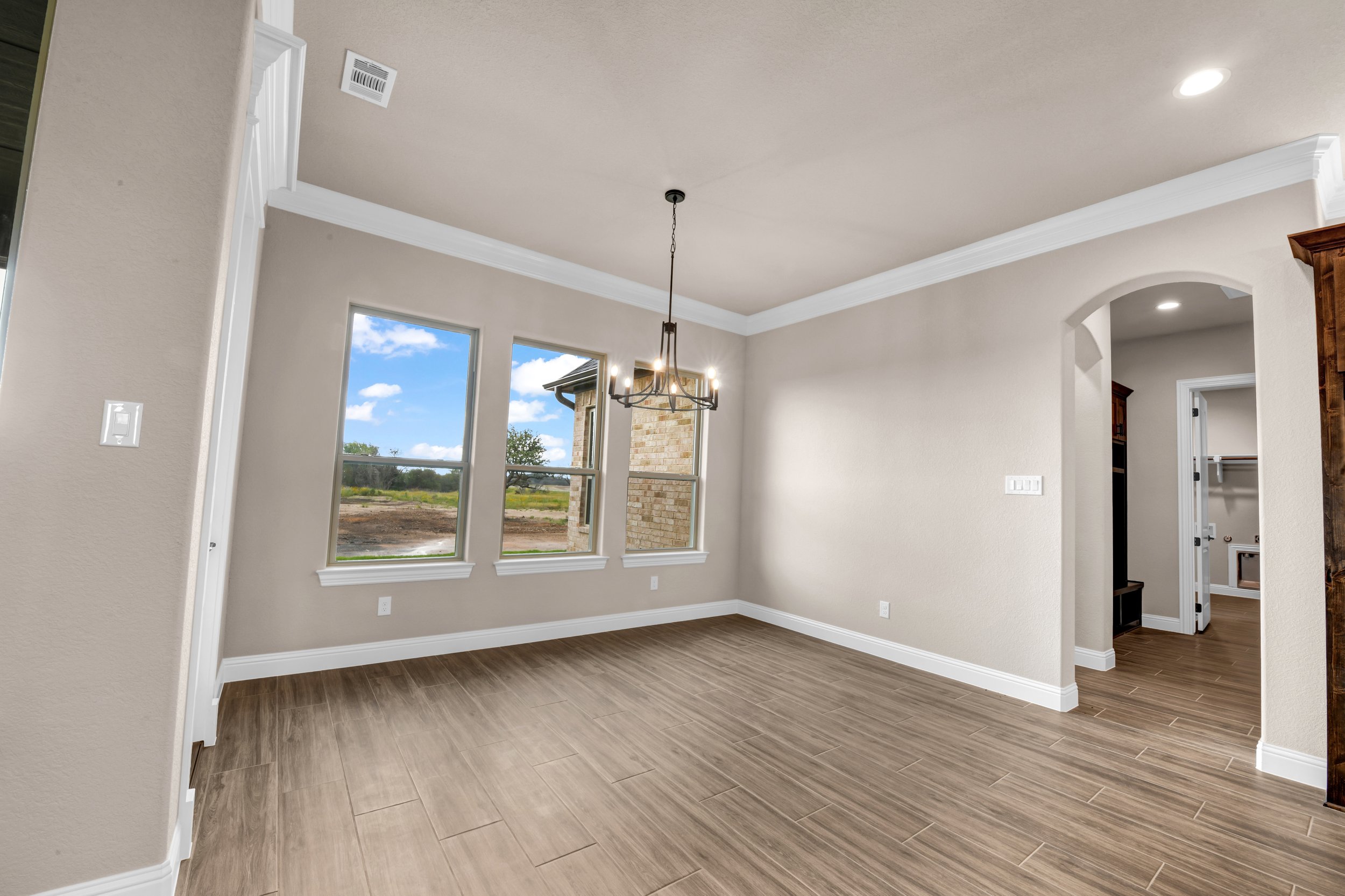Empty living room with wood-look tile flooring, beige walls, white crown molding, three large windows, a chandelier, and an arched doorway leading to another room.