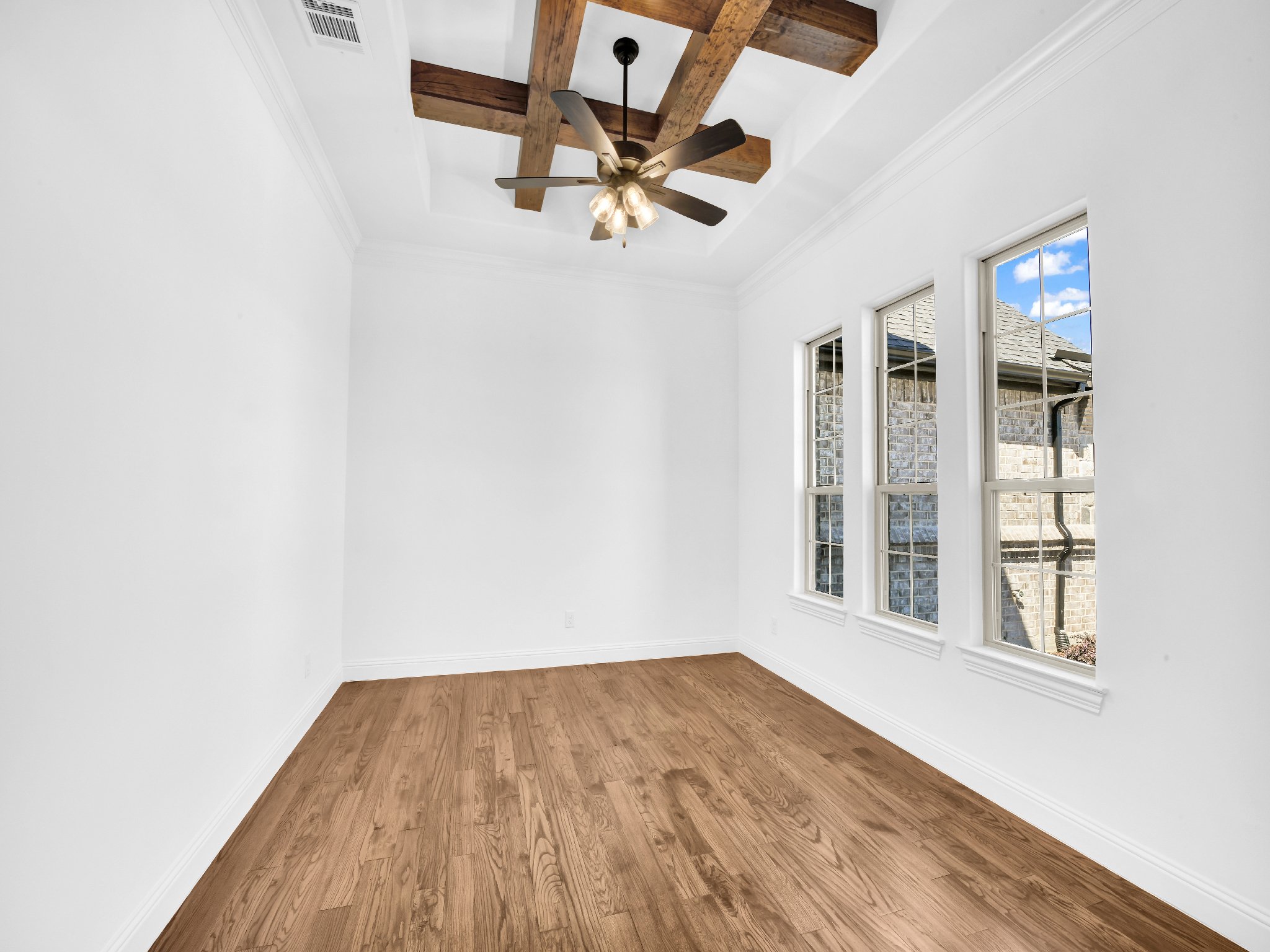 Empty room with white walls, hardwood floor, and three large windows showing an exterior brick wall and blue sky