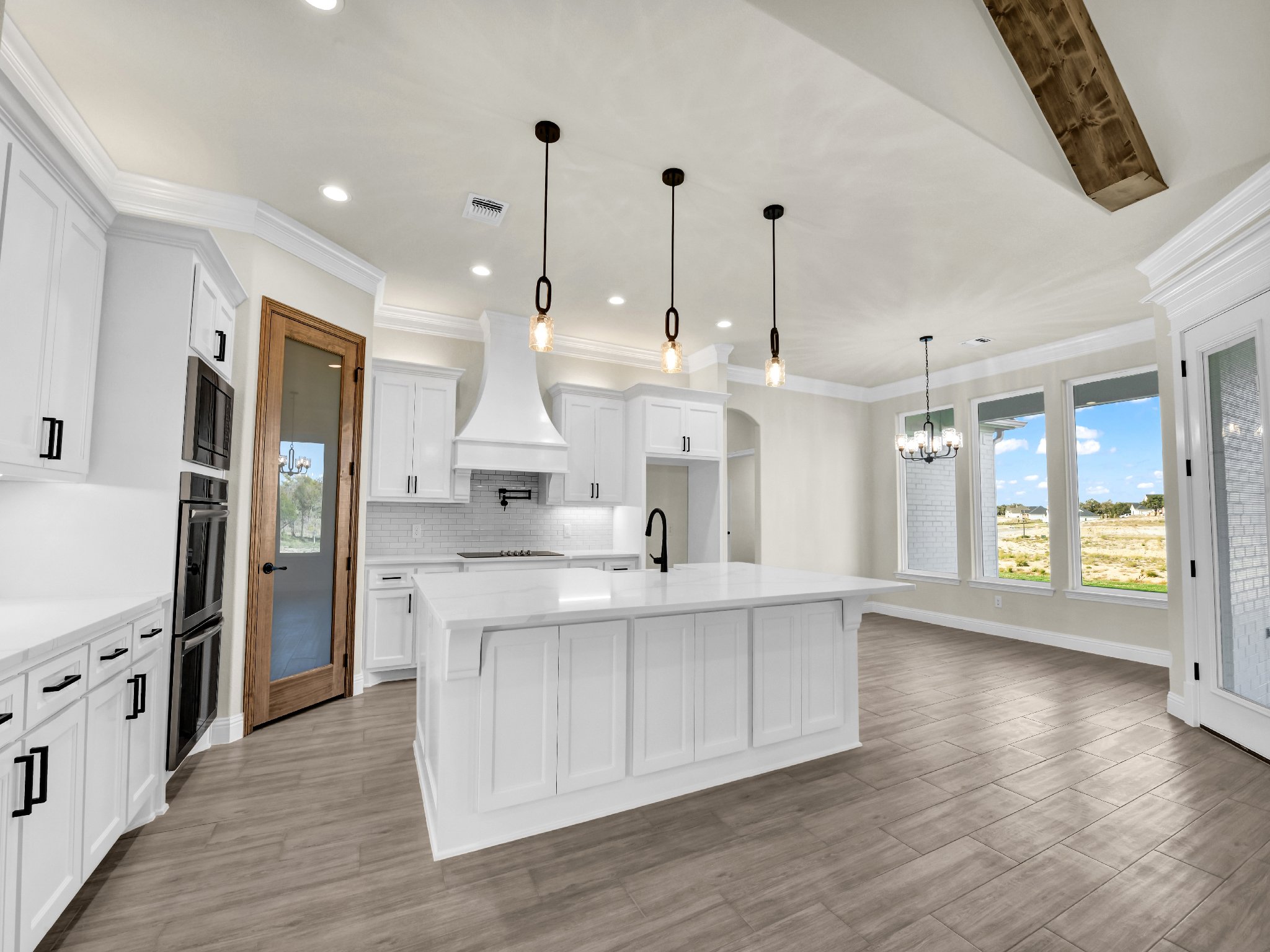 Modern kitchen with white cabinets, a large island, black fixtures, wood accents, and large windows overlooking a landscape.
