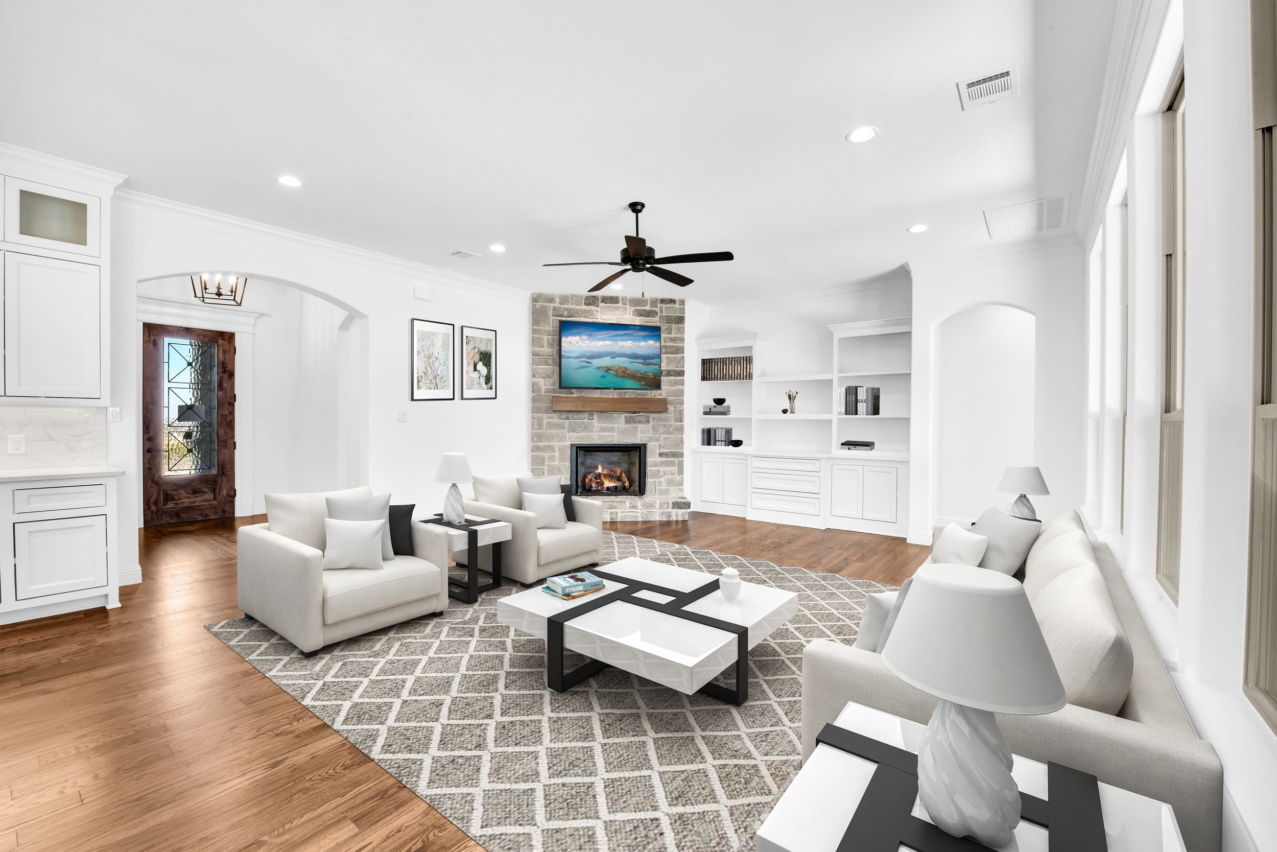 Bright living room with white couches, a stone fireplace, a wall-mounted TV, wooden flooring, built-in white shelves, and windows with natural light.