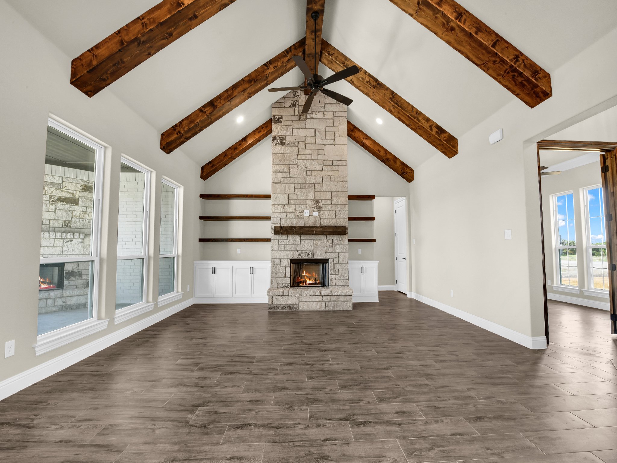 Living room with vaulted ceiling, exposed wooden beams, stone fireplace, built-in white cabinets, hardwood floors, and large windows.