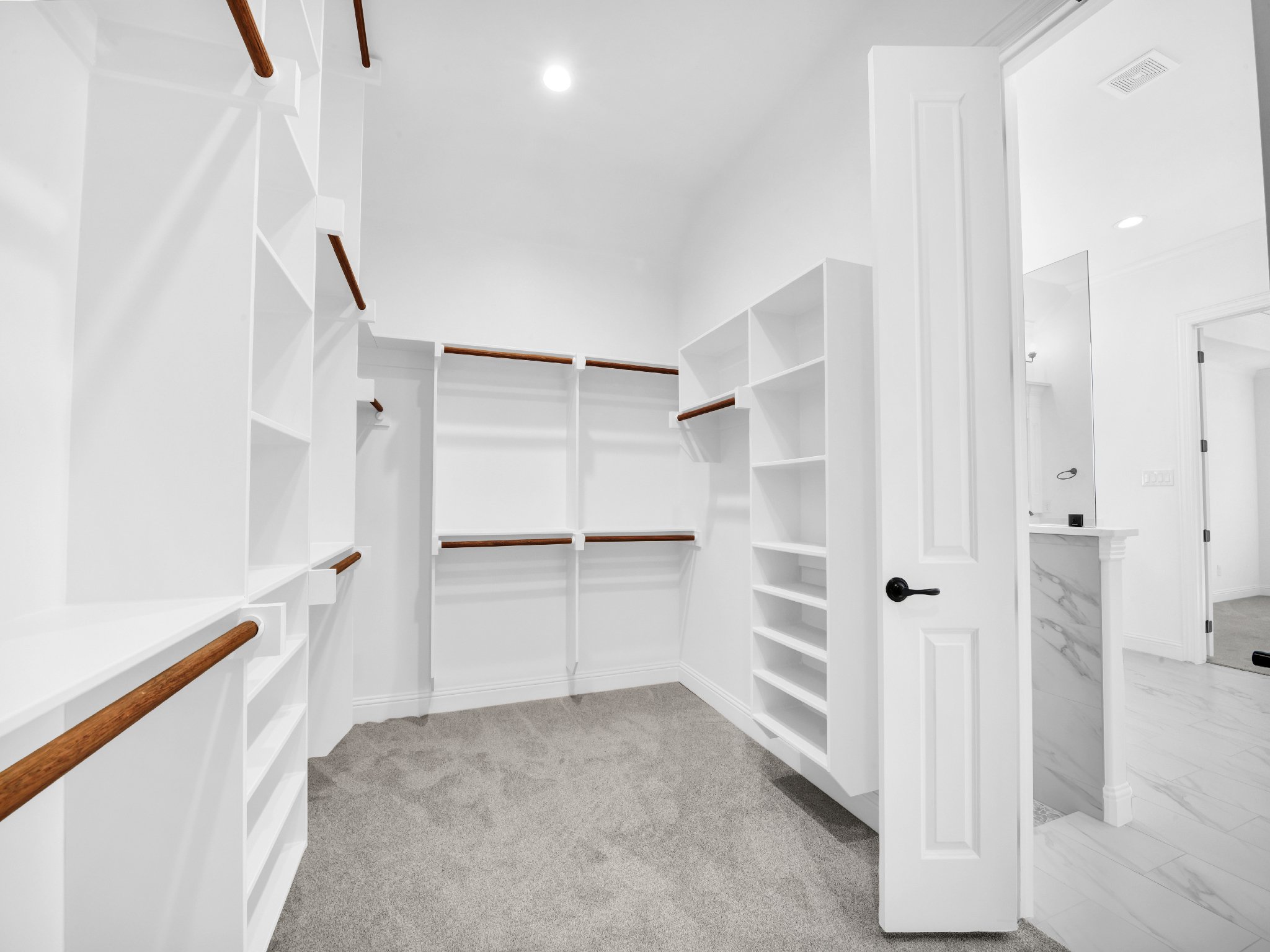 Empty walk-in closet with white shelves, wooden rods, gray carpet, and a door leading to a bathroom with marble-style tiles.