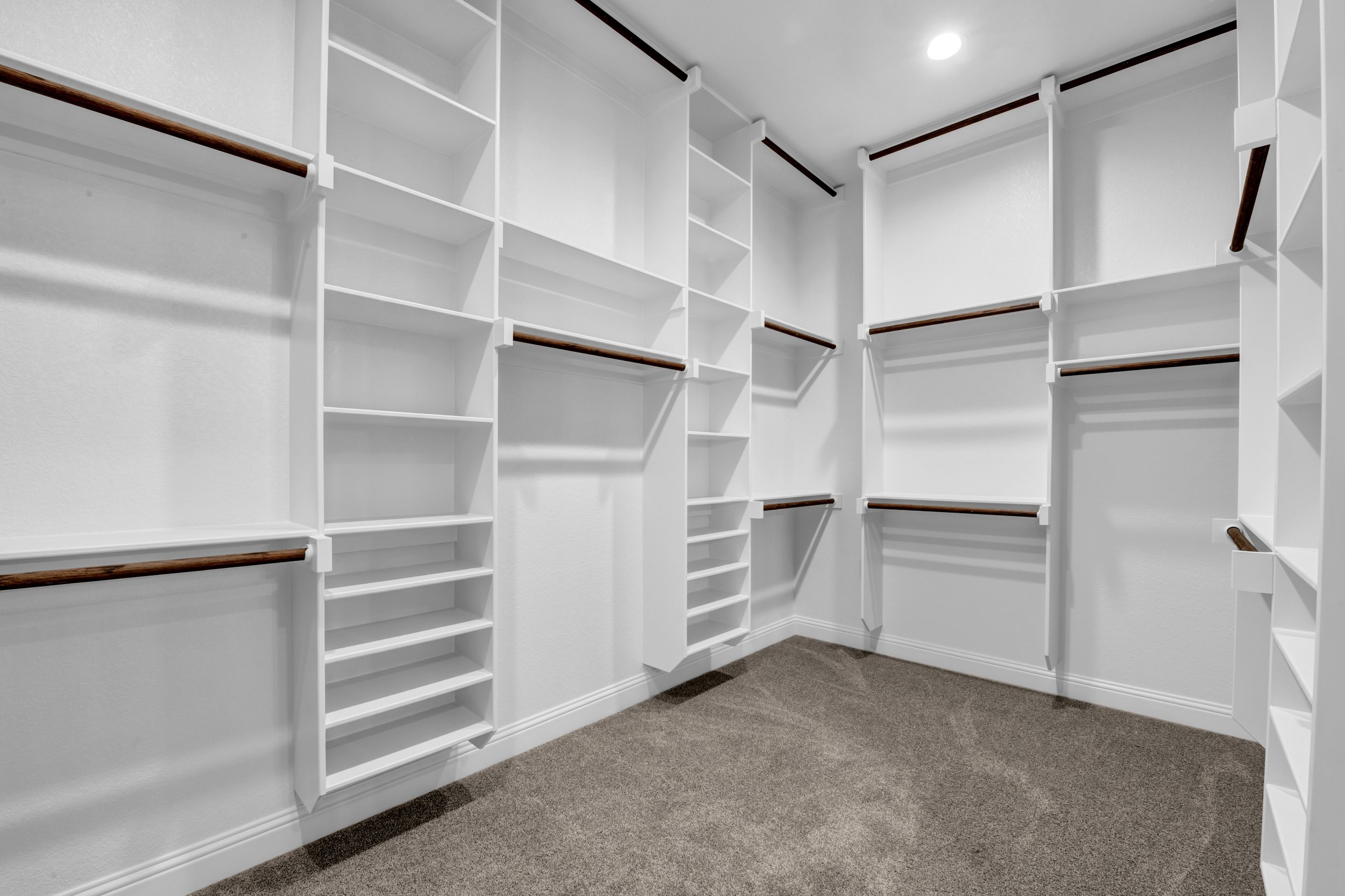 Empty walk-in closet with white shelves and wooden hanging rods, beige carpet floor, and recessed ceiling light