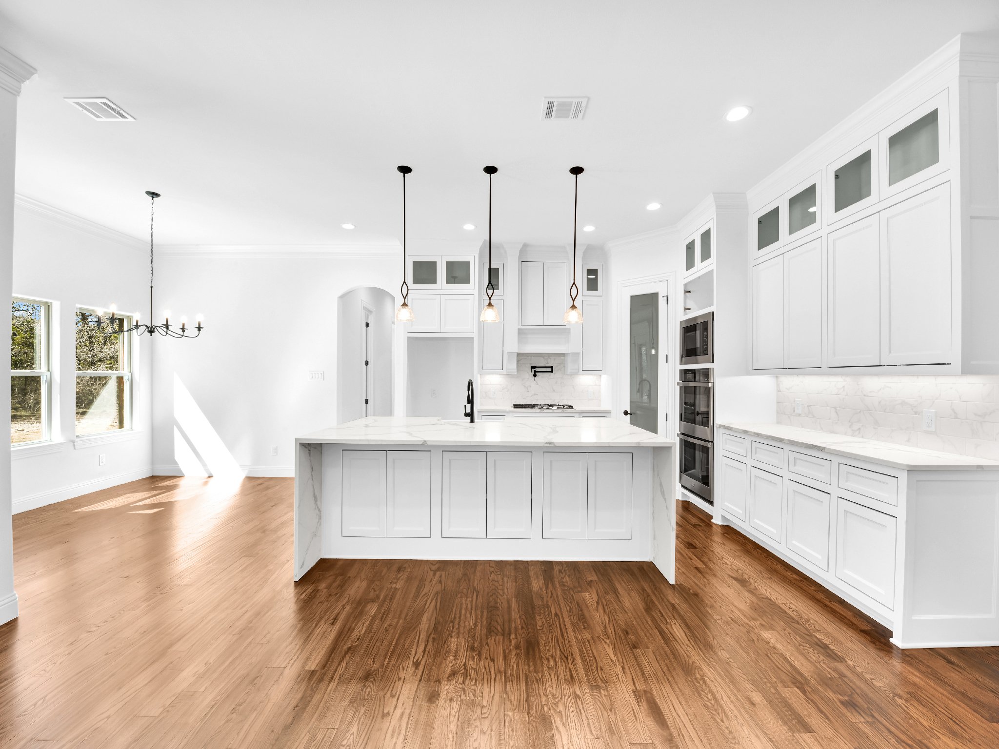 Modern white kitchen with marble countertops, wooden flooring, and pendant lighting over island, adjacent to dining area with chandelier, large windows providing natural light.