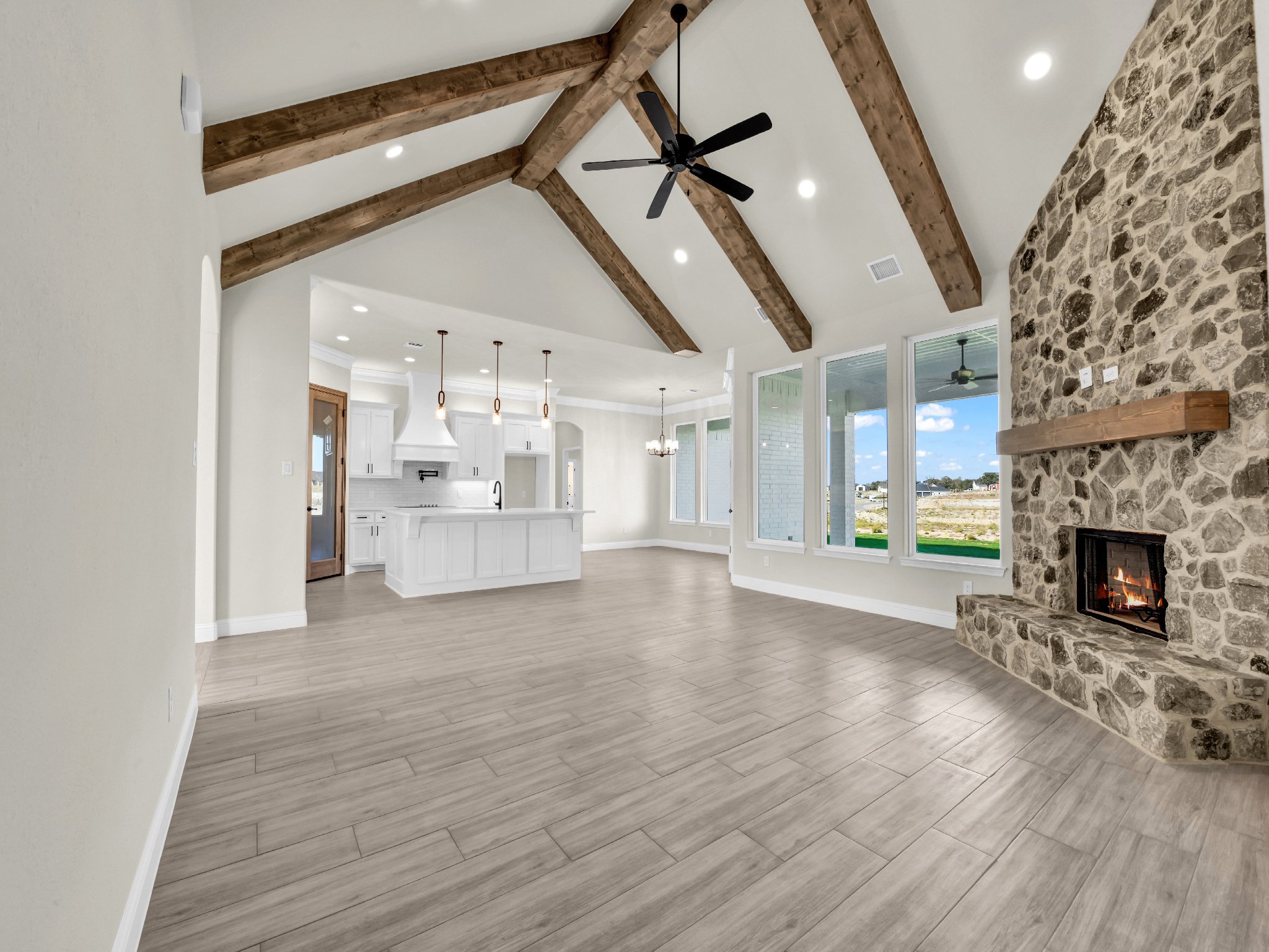 Open living area with vaulted ceiling, exposed wooden beams, a black ceiling fan, a stone fireplace, large windows, and light wood flooring, leading to a white kitchen with an island, pendant lights, and a chandelier.