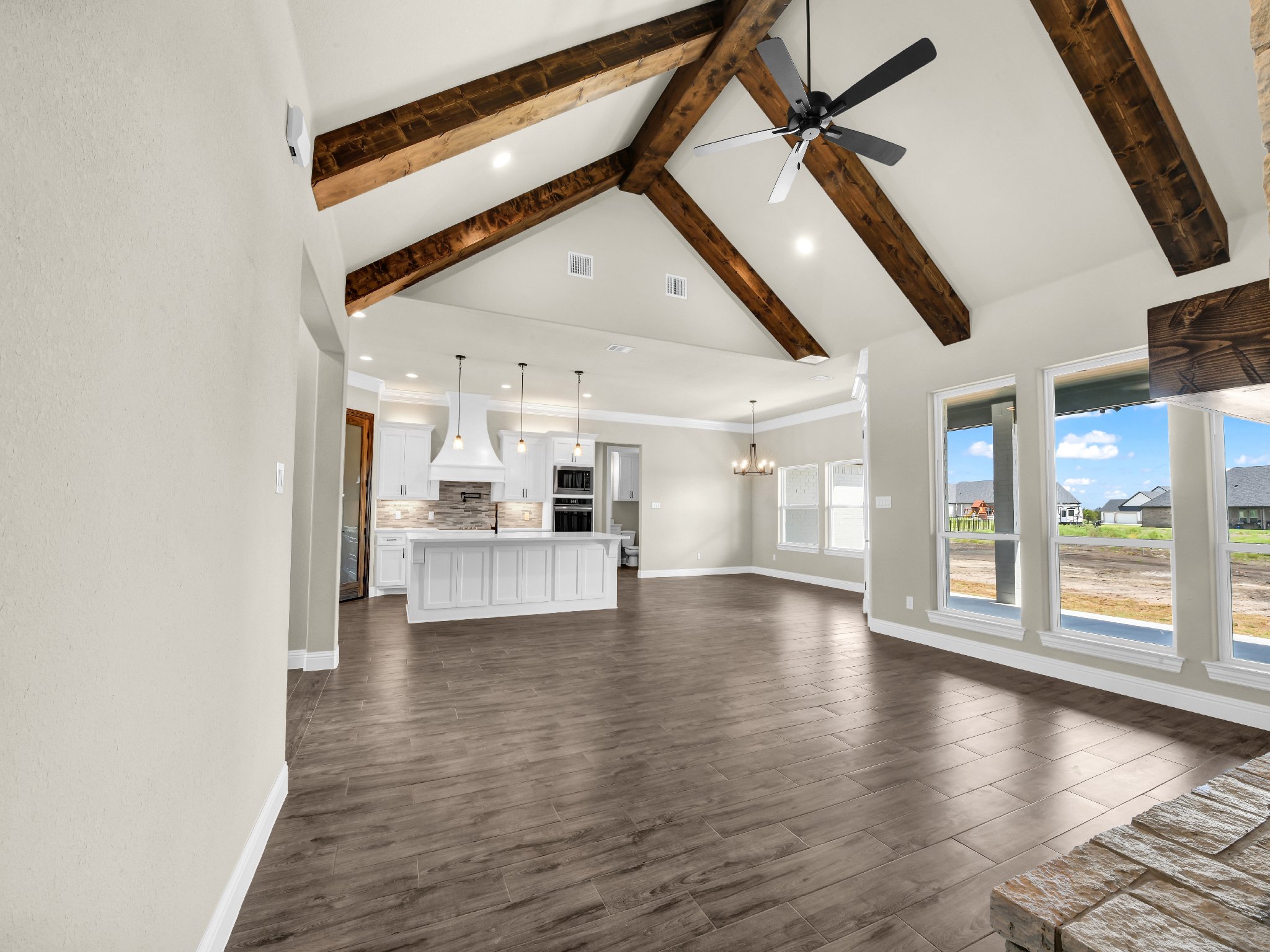 Empty open-concept living and kitchen area with high vaulted ceiling, exposed wooden beams, and large windows showing an outdoor view.