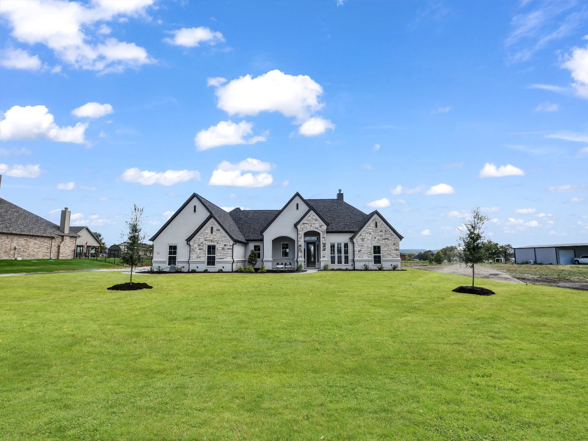 A modern white house with stone accents on a large grassy lawn with two small trees and a driveway. Blue sky with clouds.