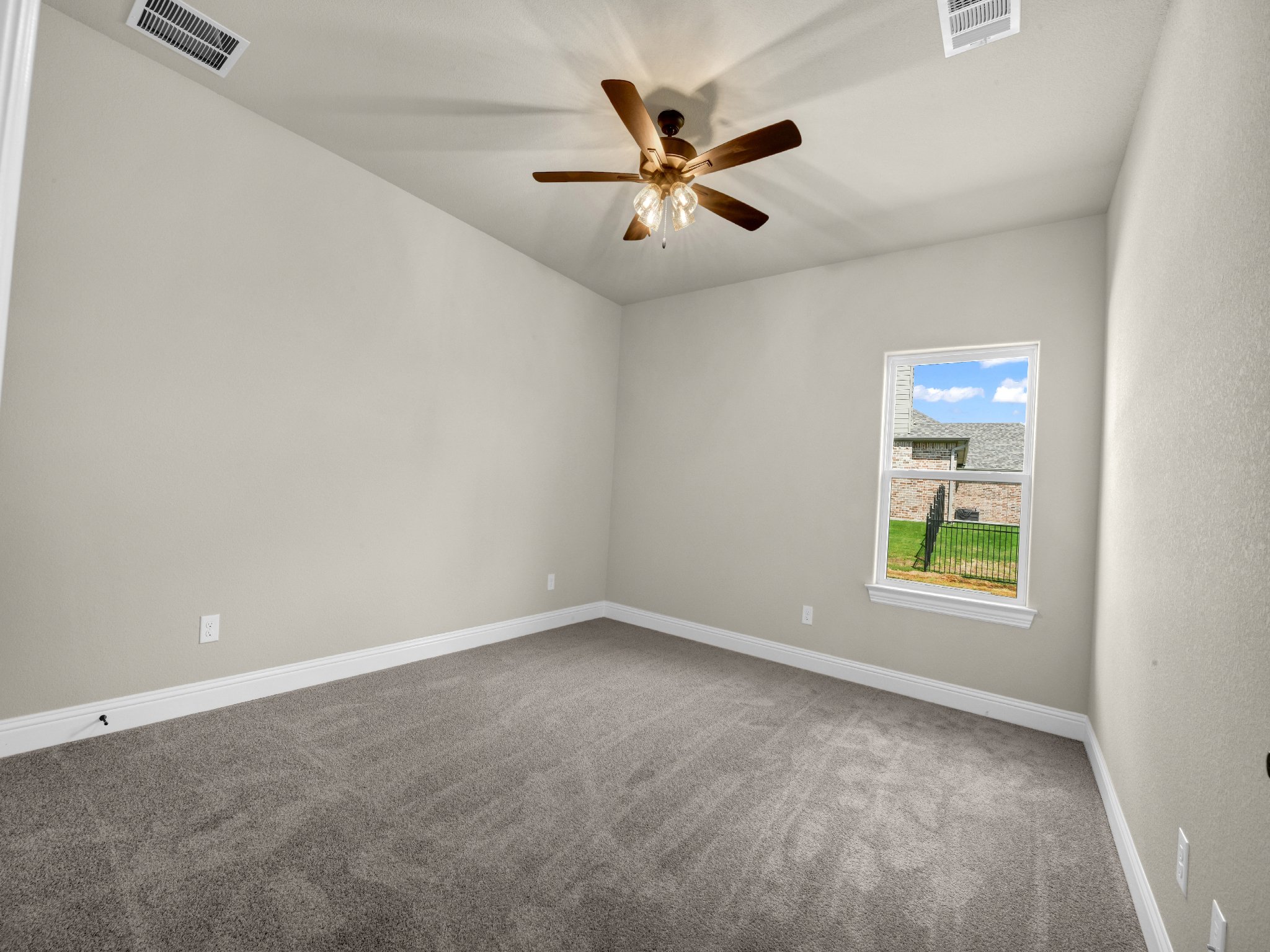 Empty room with beige walls, gray carpet, white baseboards, a window showing a backyard, and a ceiling fan with wooden blades.