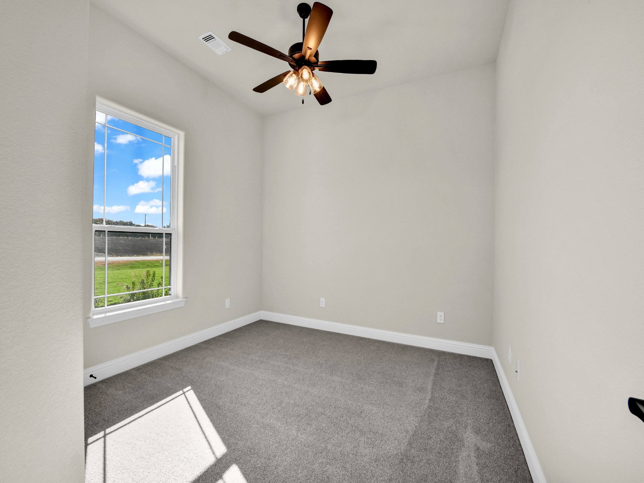 Empty white room with beige carpet, a window showing blue sky and clouds, a ceiling fan with lights, and multiple electrical outlets.