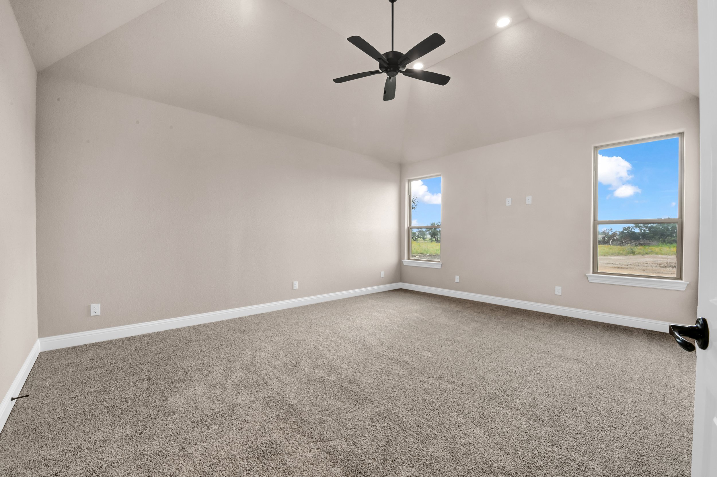 Empty bedroom with beige walls, beige carpet, two windows showing a blue sky with clouds, a black ceiling fan, and white trim around the windows and baseboards.