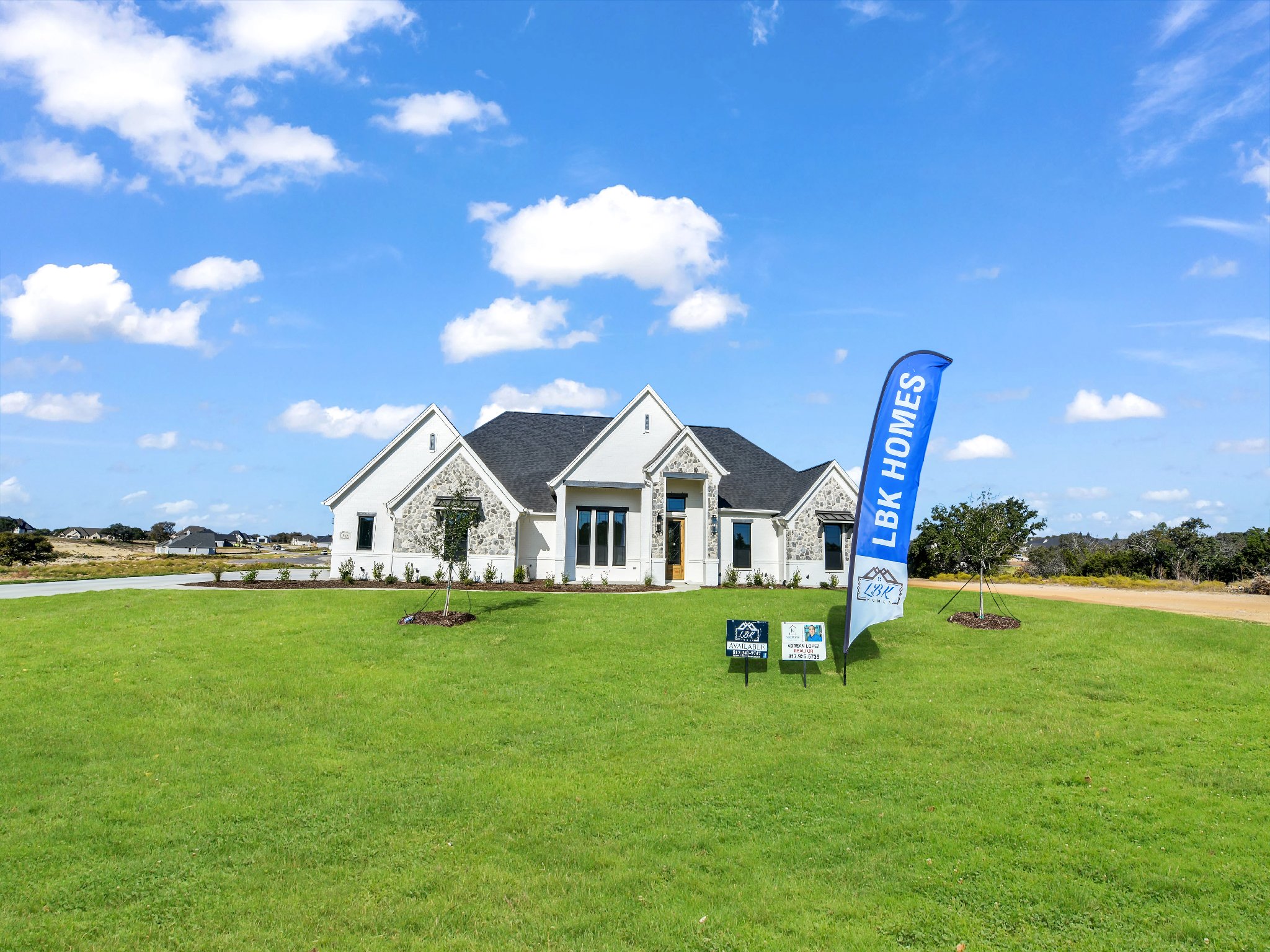 A new house with white walls and gray stone accents on a large green lawn under a partly cloudy blue sky, with a blue LBK Homes flag and signs out front.