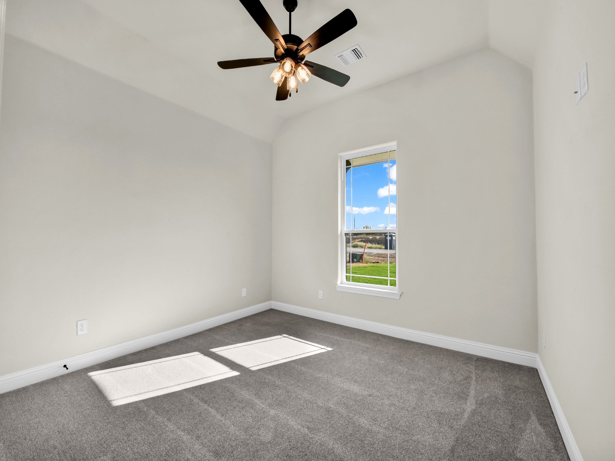 Empty bedroom with beige walls, gray carpet, a window showing a green lawn and blue sky, and a ceiling fan with lights