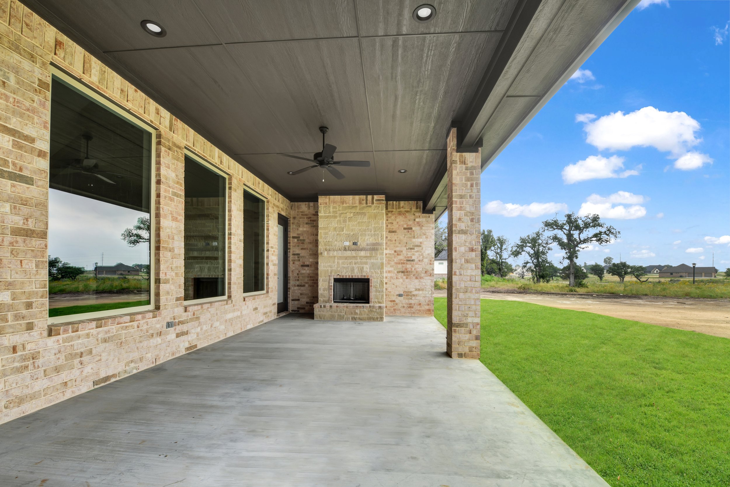 Covered outdoor patio area with brick walls, concrete floor, ceiling fan, and fireplace, overlooking a grassy yard and open land with trees and blue sky.