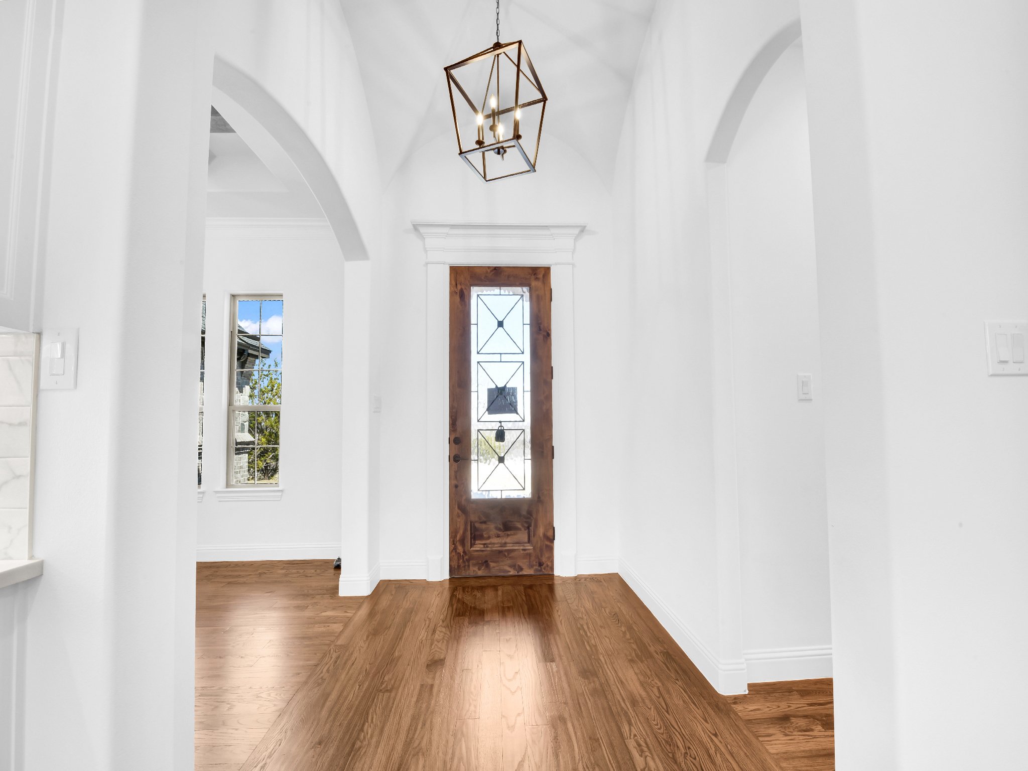 Empty entryway with wooden door, white walls, hardwood floor, and a modern geometric chandelier hanging from the ceiling.