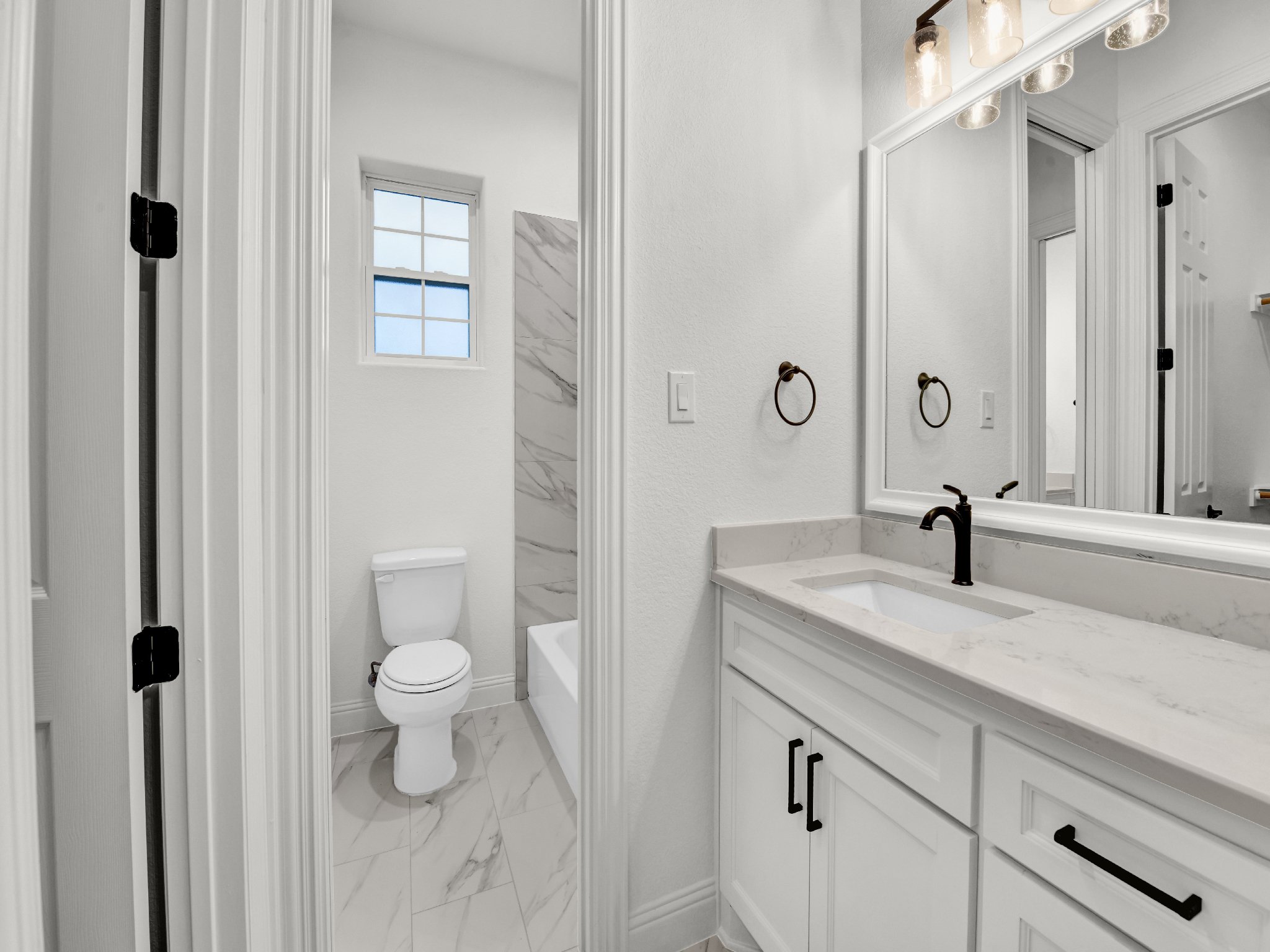 Modern white bathroom with a vanity, rectangular sink, black fixtures, marble countertop, mirror, towel rings, and a separate toilet and shower area with a window.