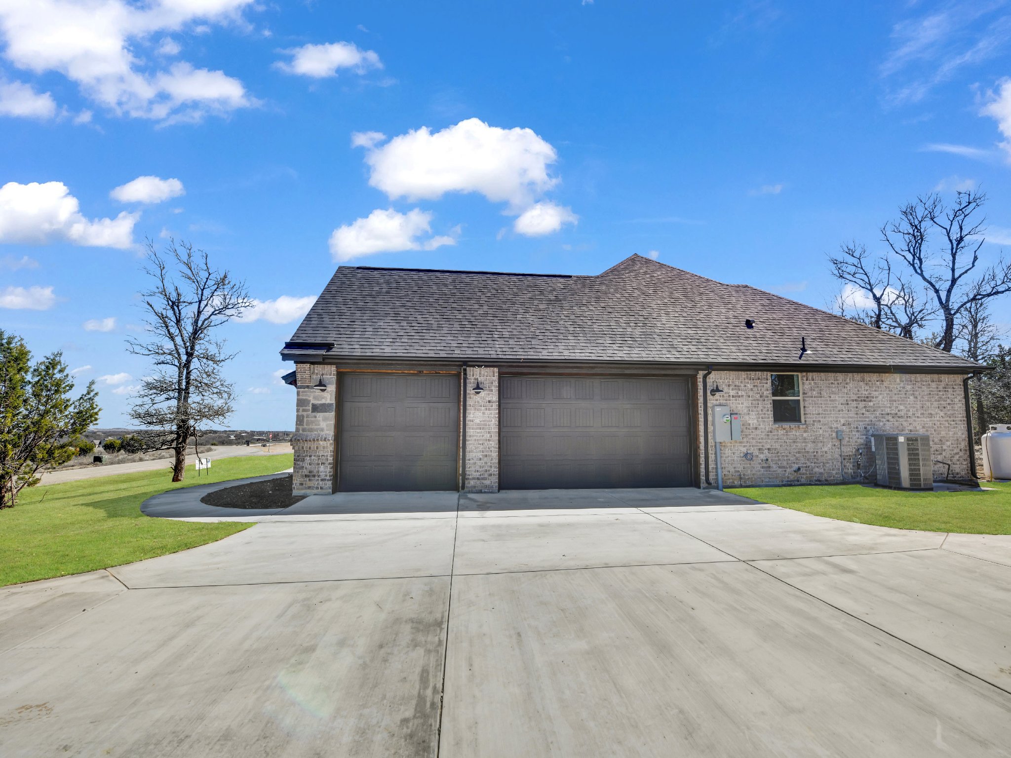 A single-story brick house with two garage doors, a sidewalk, and green lawn under a blue sky with clouds.
