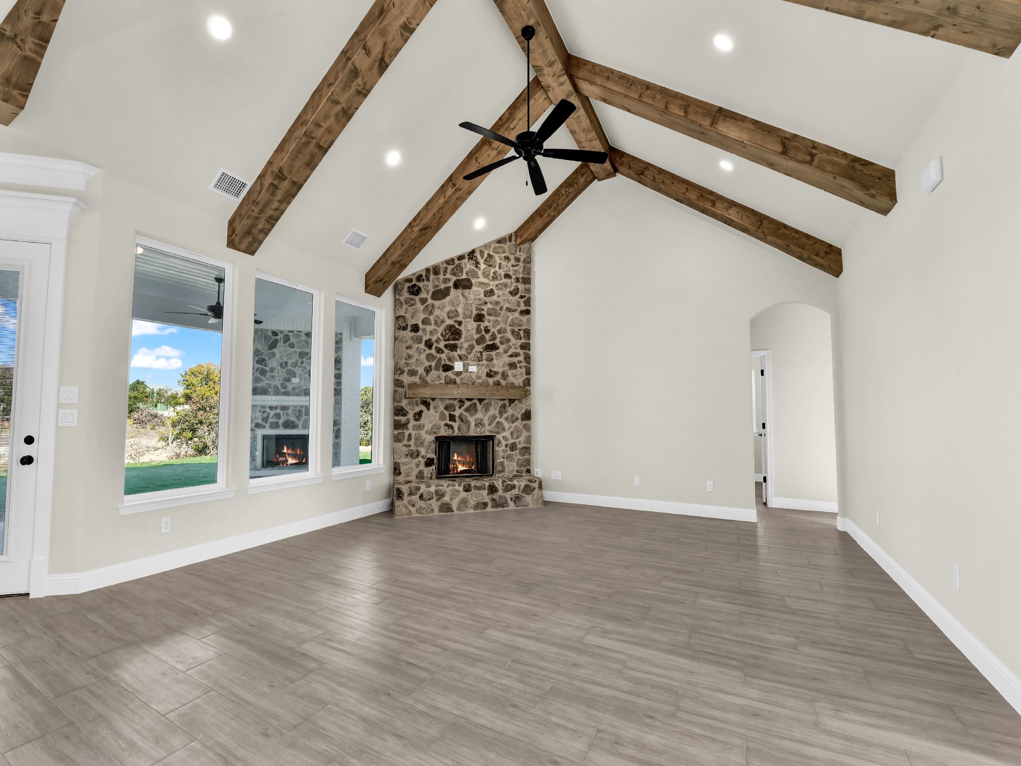Empty living room with high vaulted ceiling featuring exposed wooden beams, a stone fireplace, large windows, a ceiling fan, and light-colored hardwood floors.