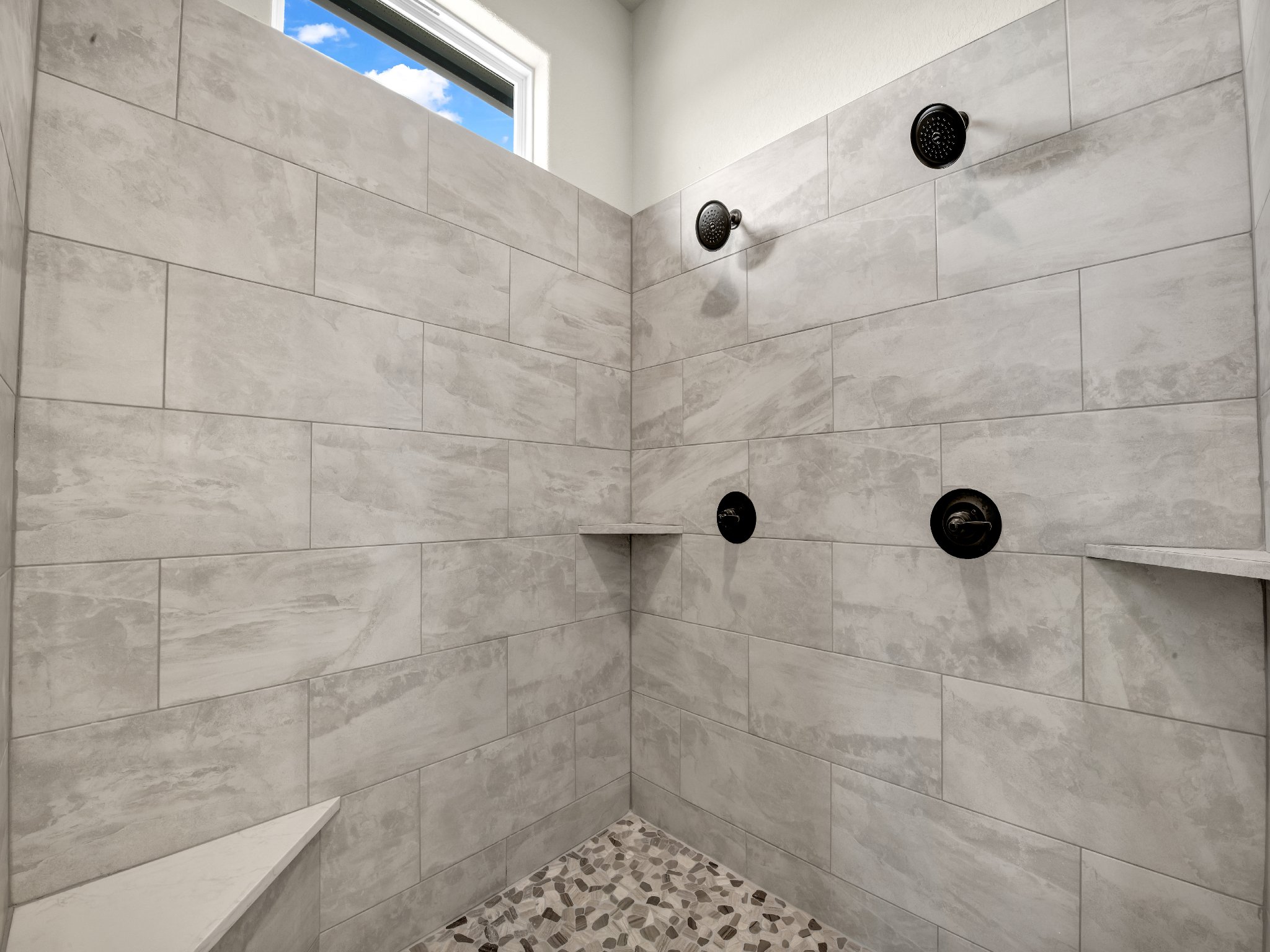 Empty walk-in shower with beige tiles, black fixtures, a small shelf, pebble floor, and a window showing blue sky and clouds.