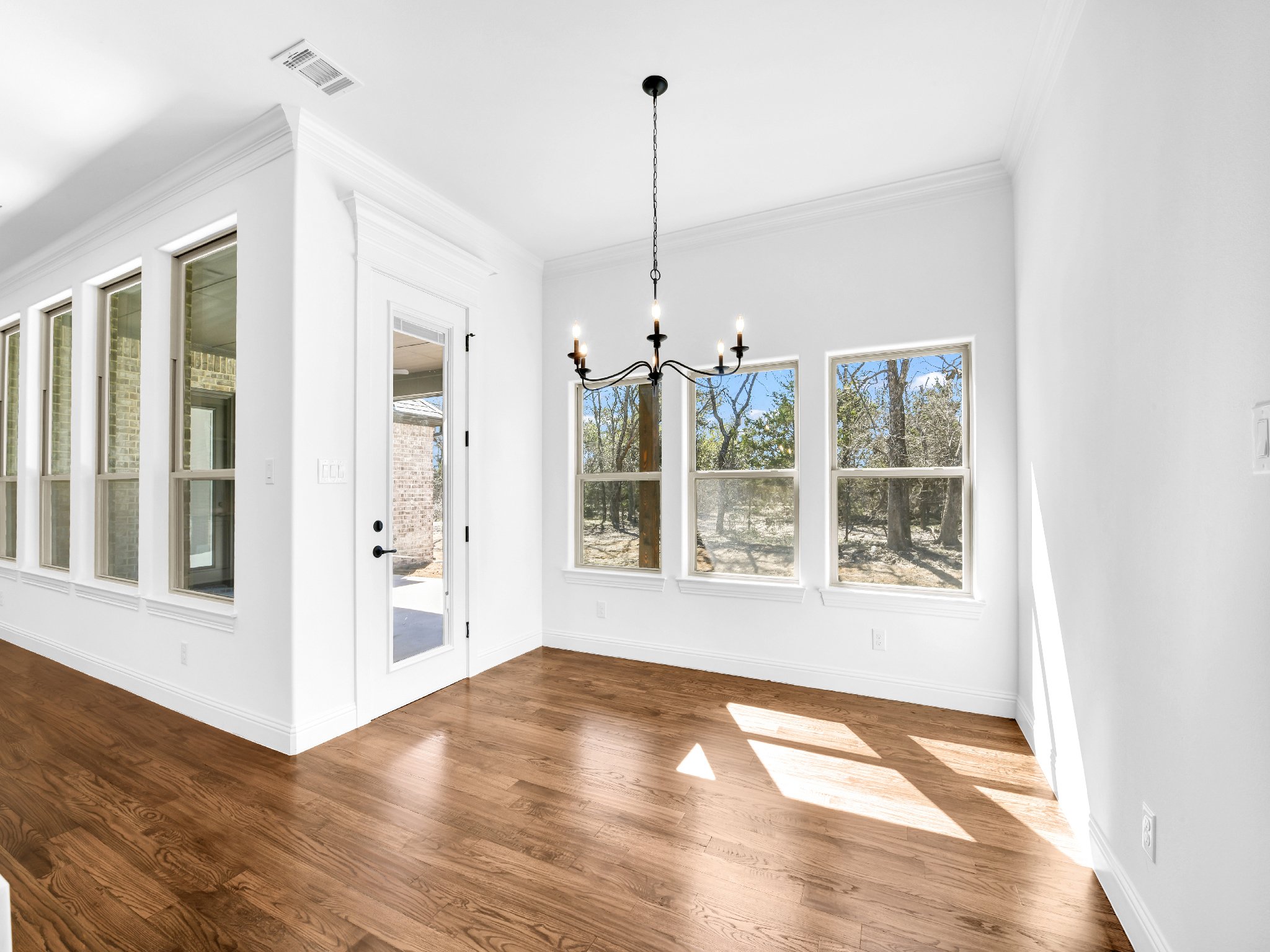 Empty room with white walls, hardwood floor, windows showing outdoor trees, and a black chandelier