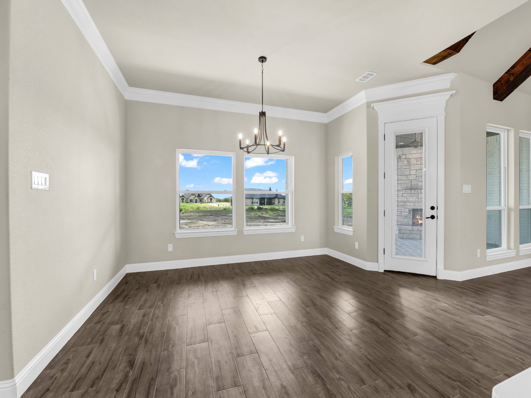 Empty room with beige walls, white crown molding, and dark wood flooring. Large windows with white trim letting in natural light, and a white door with glass leading outside. Ceiling with a chandelier and wooden beams.