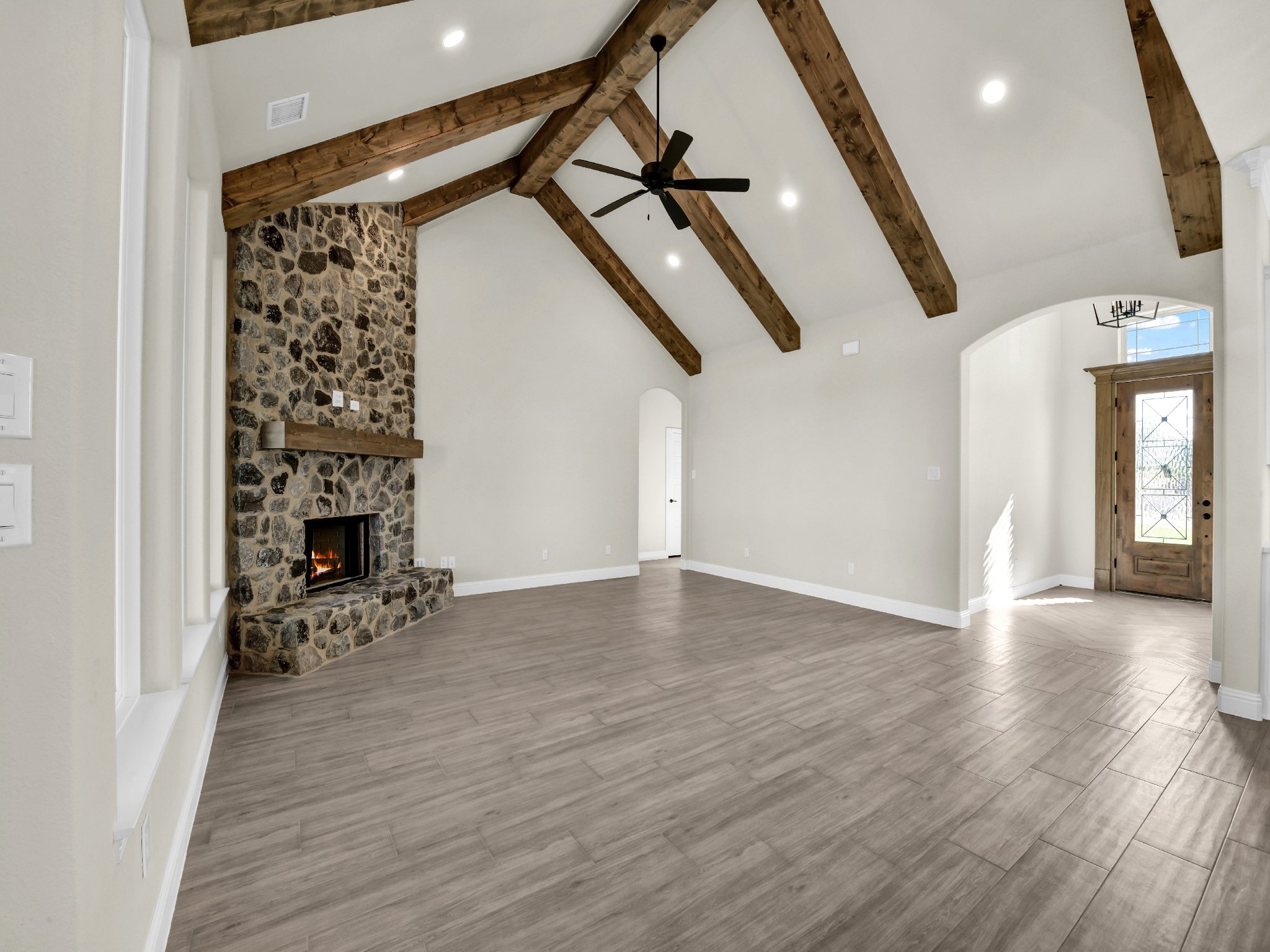 Empty living room with wood plank floors, stone fireplace, wooden exposed beam ceiling, ceiling fan, and front door.